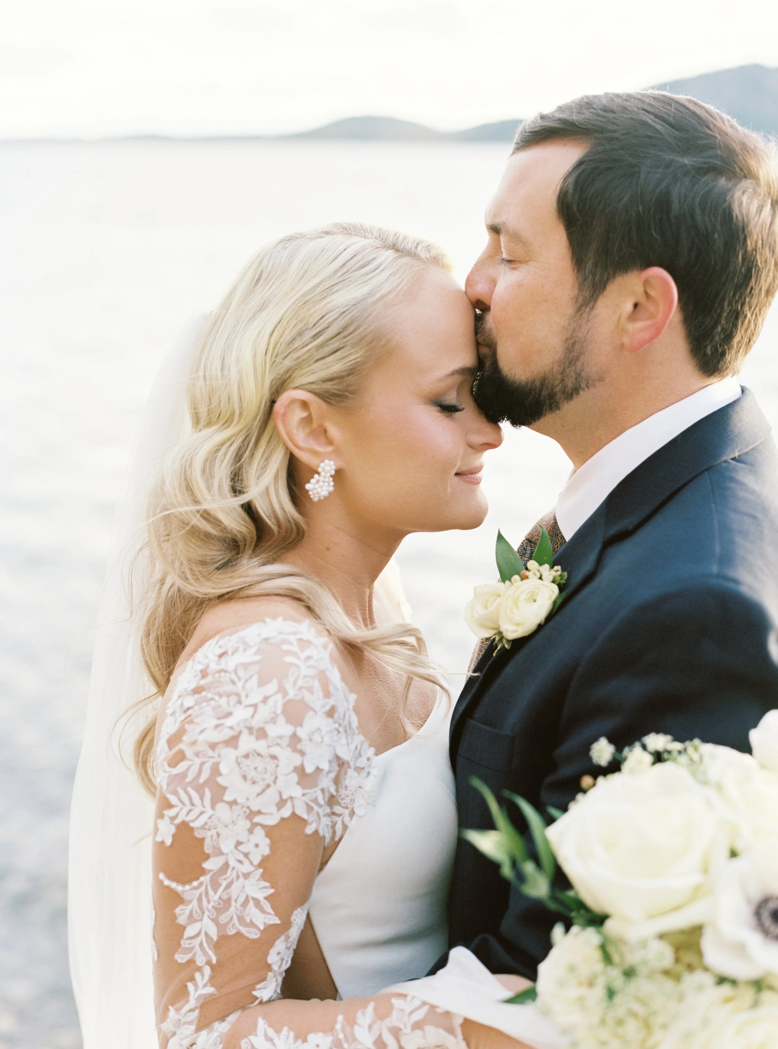 A bride and groom share a kiss, with the groom giving a gentle kiss to the bride's forehead, standing outdoors near a body of water with hills in the background.