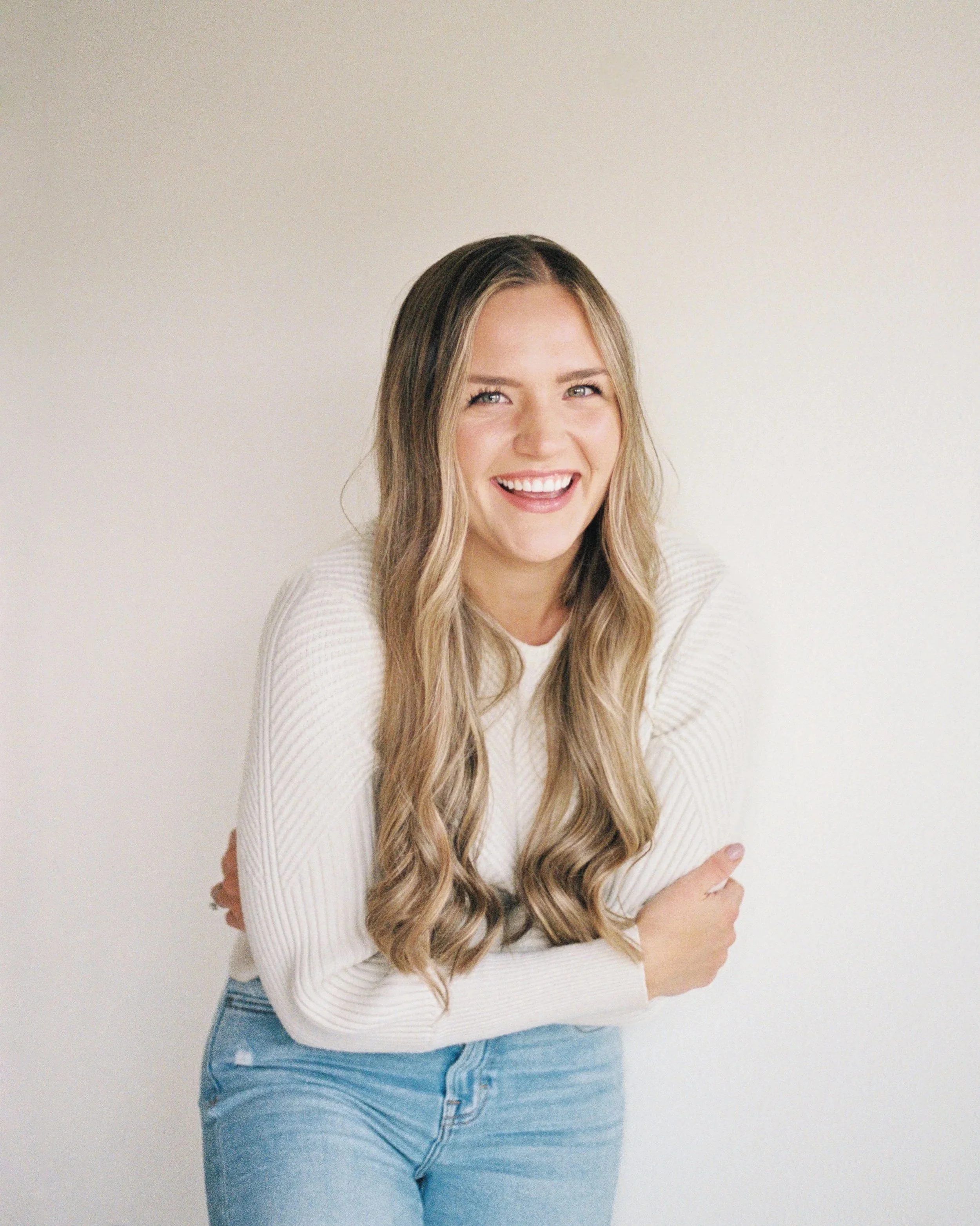 A young woman with long wavy hair smiling in front of a plain white wall.