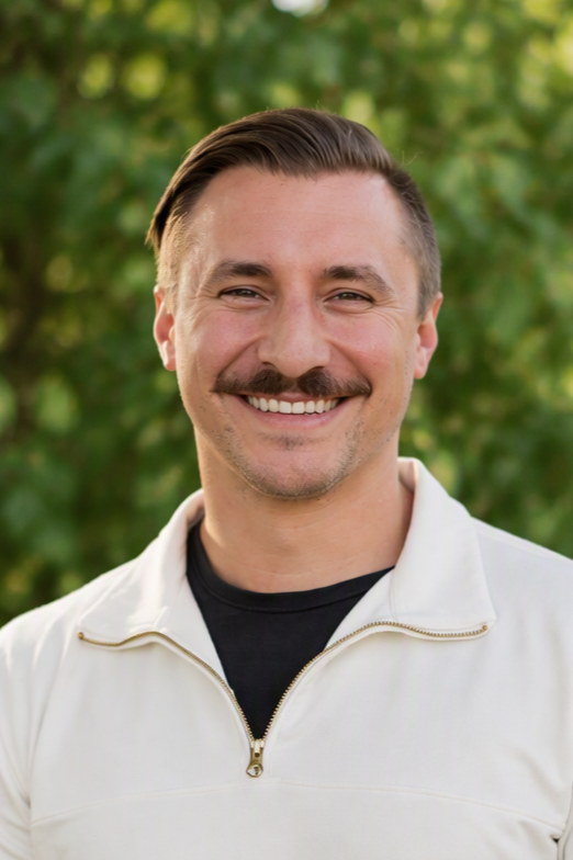 A smiling man with a mustache and styled hair stands outdoors with snow-capped mountains and a cityscape in the background.