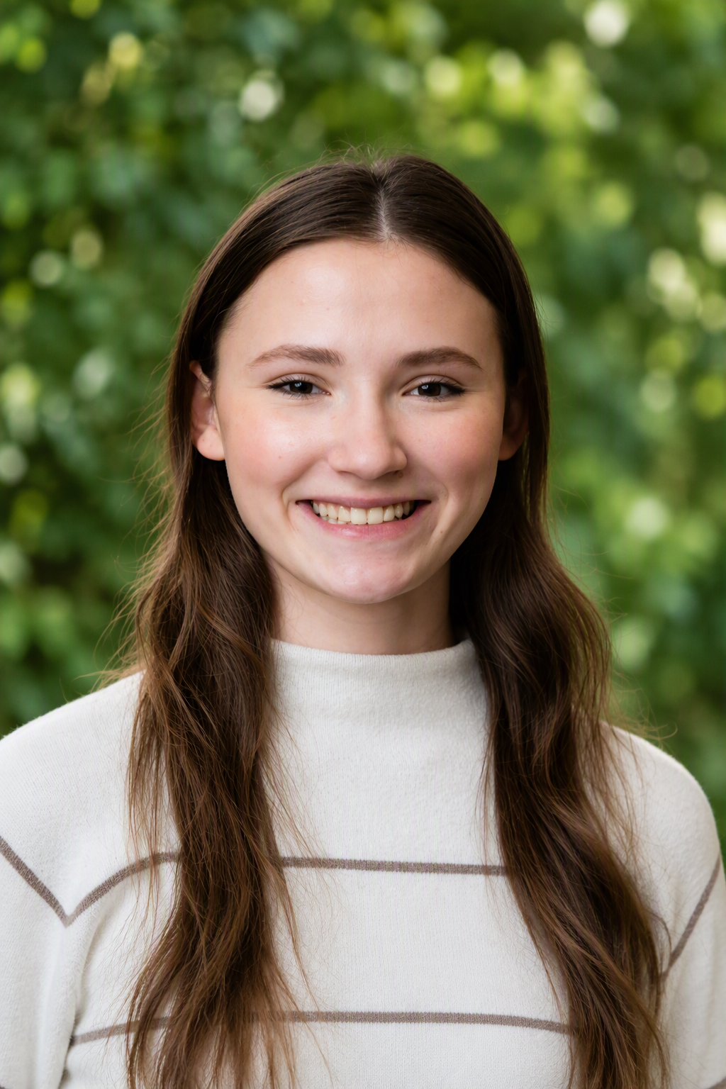 A young woman with long brown hair and a white top, smiling with a natural background.