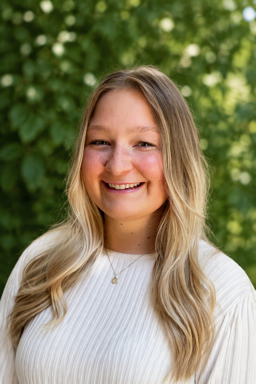 A young woman with long wavy blonde hair smiling in front a leafy background