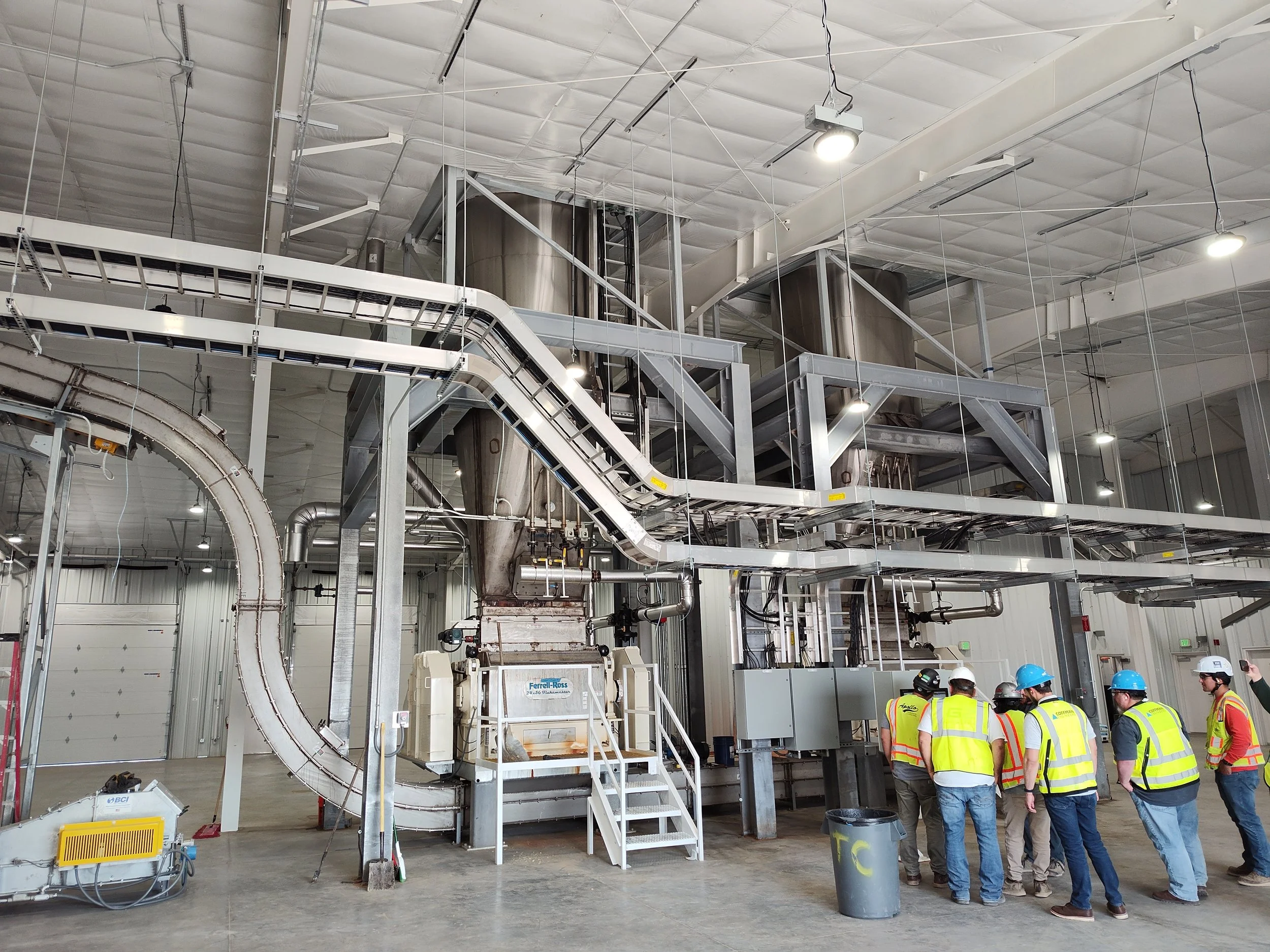 Group of workers in safety vests and helmets touring an industrial facility with large machinery and metal piping.
