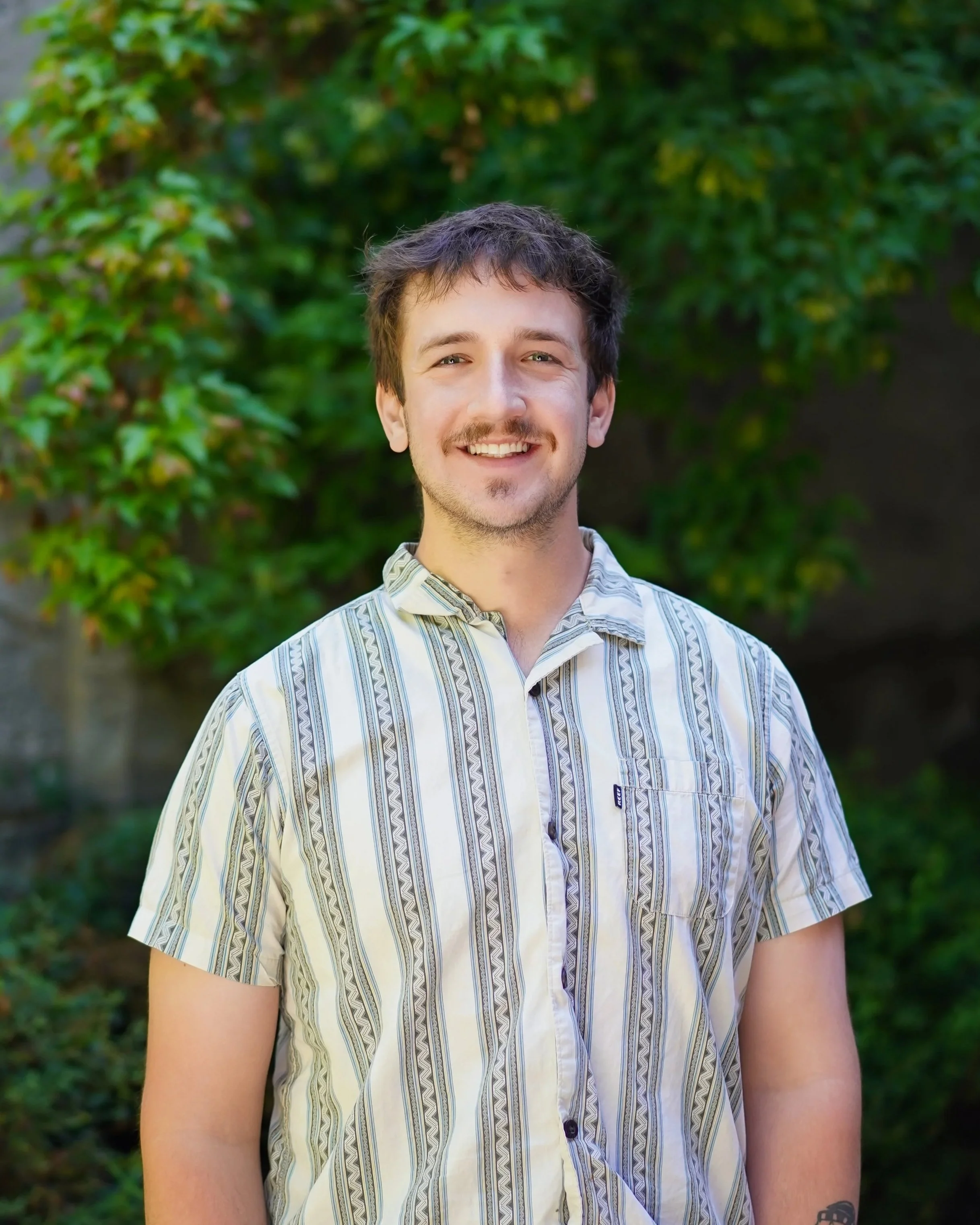 A man standing outdoors with a background of green foliage, smiling at the camera, wearing a short-sleeved, patterned button-up shirt.