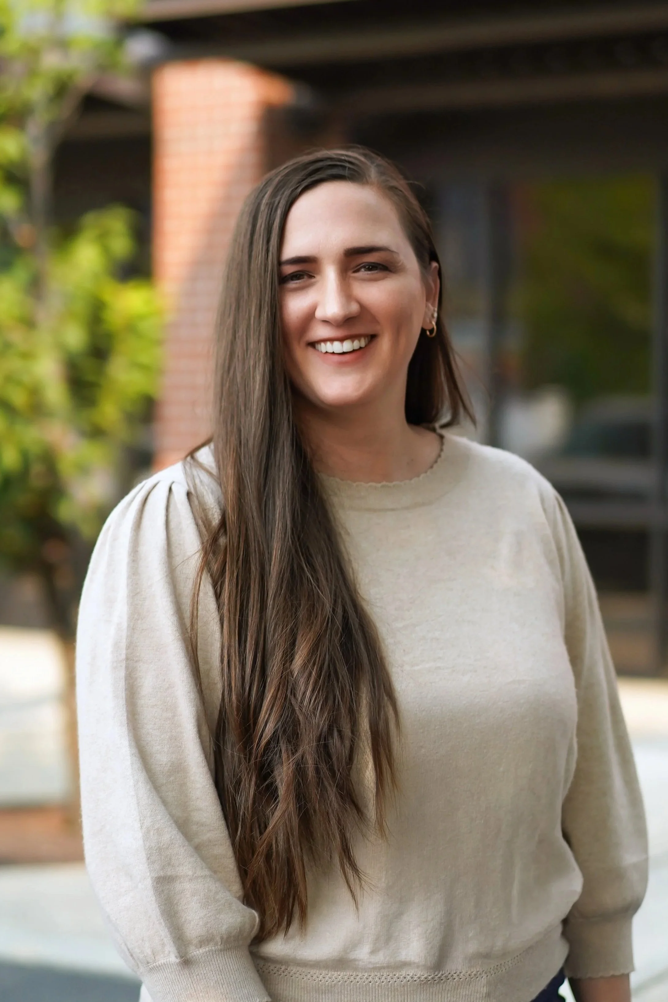 A woman with long brown hair and a bright smile, dressed in a navy blazer and white top, standing outdoors with autumn-colored trees and a building in the background.