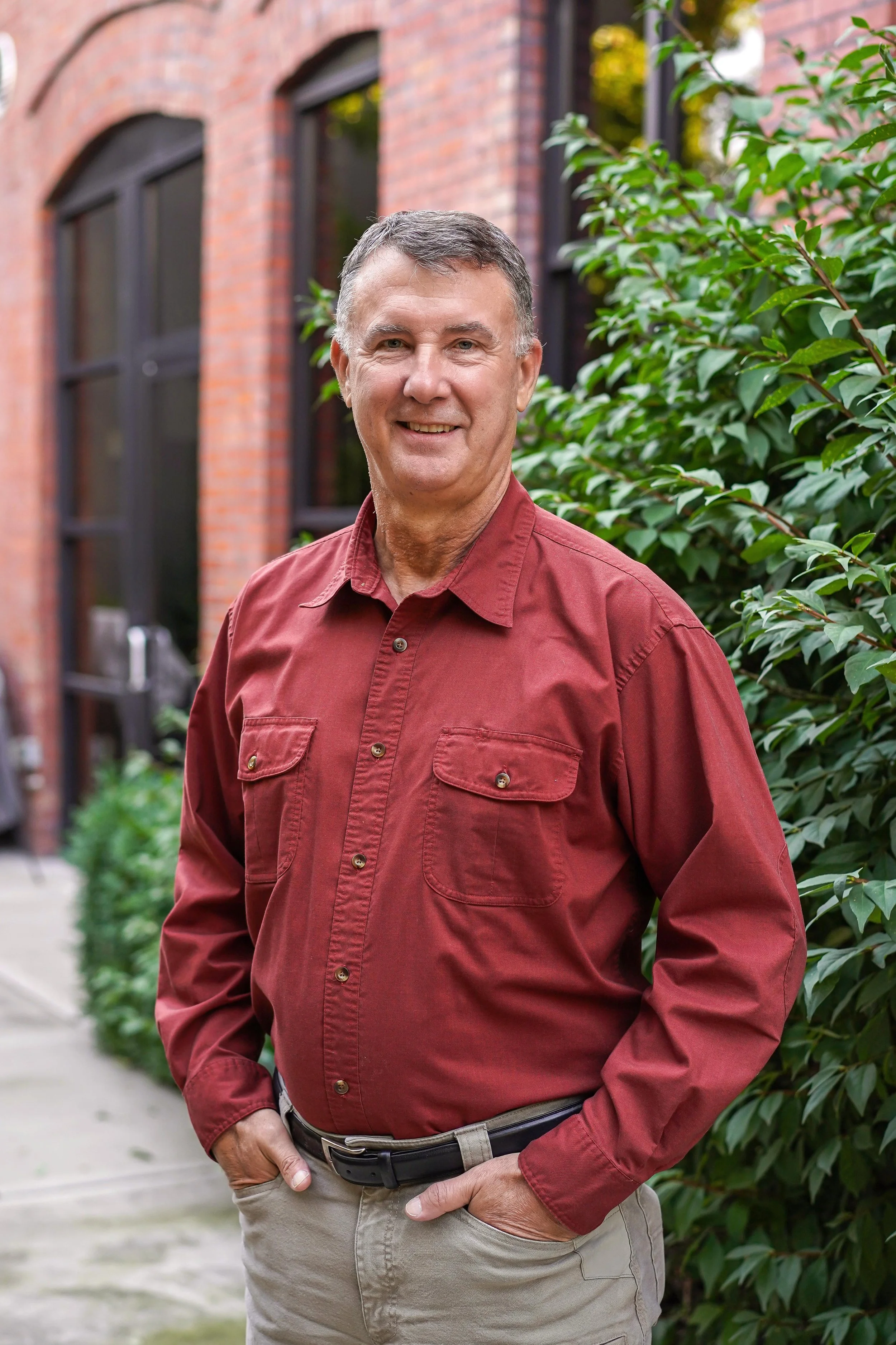 A smiling man standing outdoors in front of green trees and bushes.