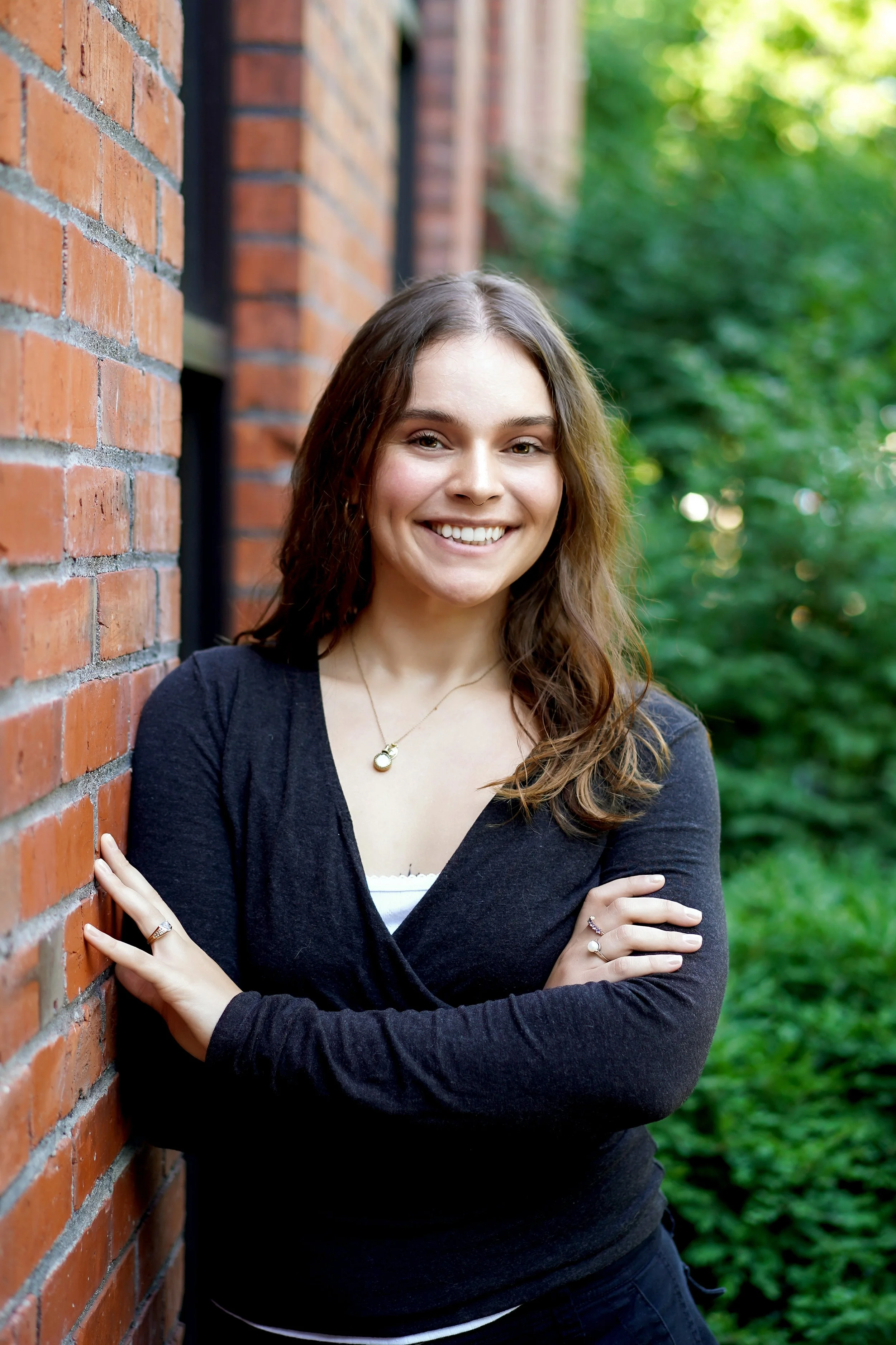 A young woman with brown hair and a smile standing outdoors, leaning against a brick wall with greenery in the background.