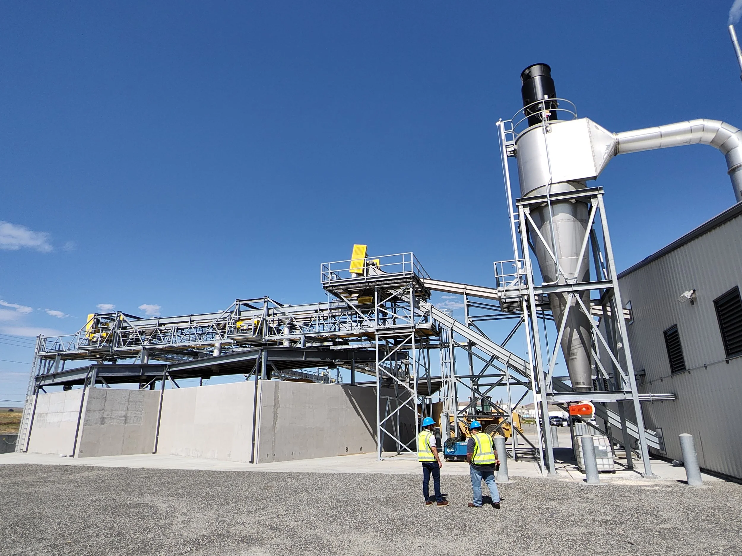 Industrial facility with metal structures, large duct, and three workers wearing safety vests and helmets standing near the building.