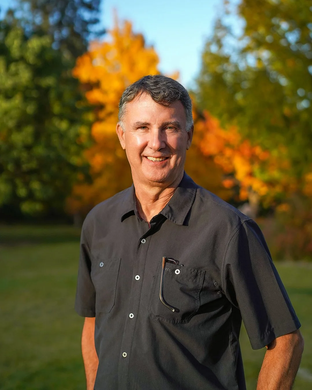 A smiling man standing outdoors in front of green trees and bushes.