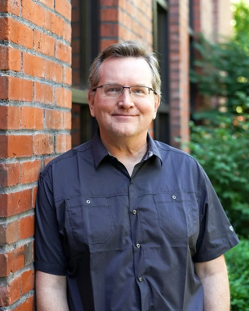 Smiling middle-aged man with glasses, wearing a navy blue shirt, standing outdoors next to a brick wall with greenery in the background.