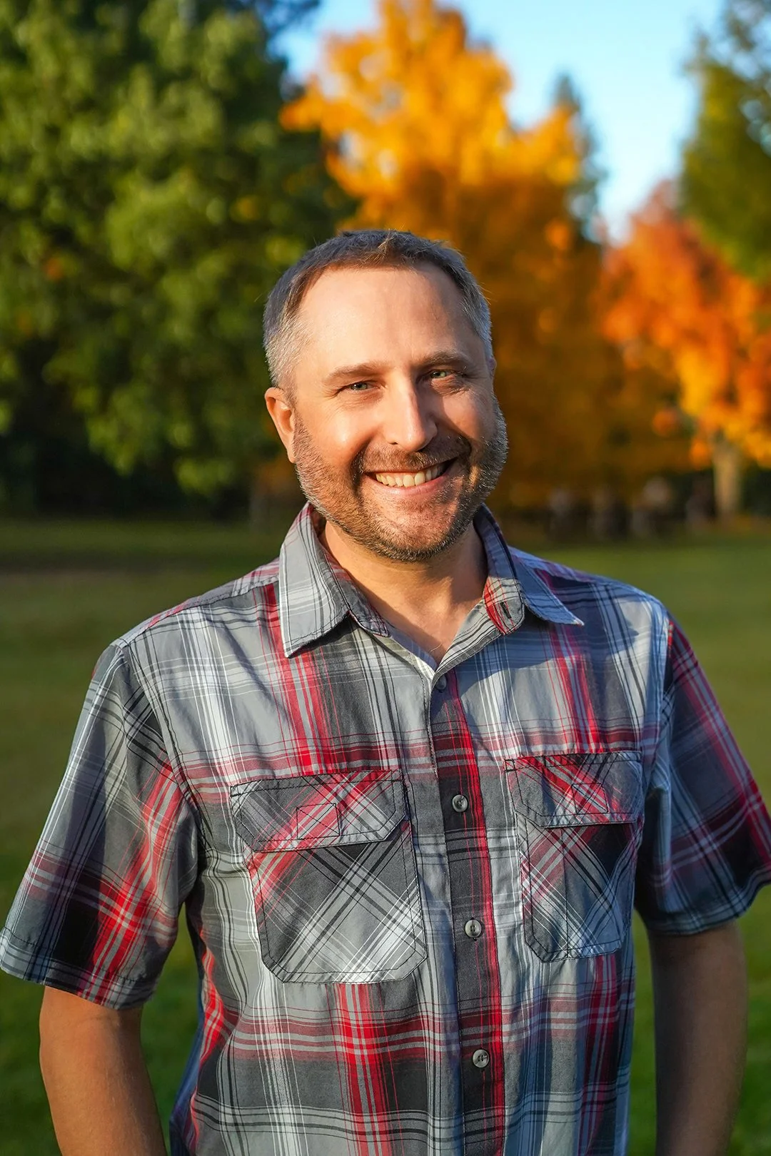 A smiling man with short, gray hair and a beard, wearing a dark plaid shirt, outdoors with green blurred trees in the background.