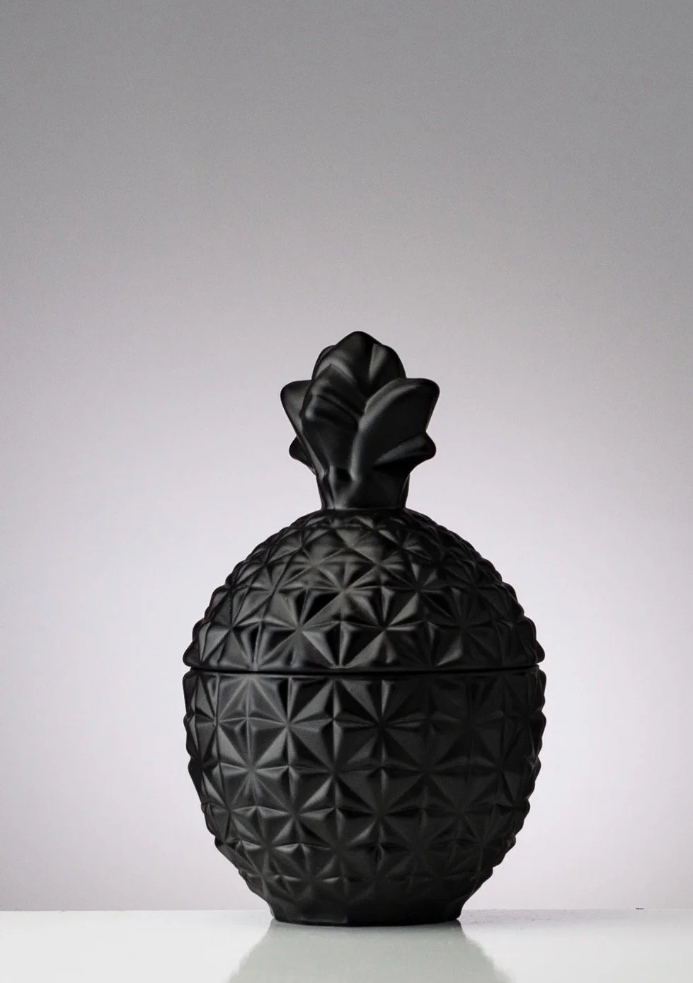 Black decorative pineapple-shaped container with textured surface and leafy top, placed on a white surface against a plain light gray background.