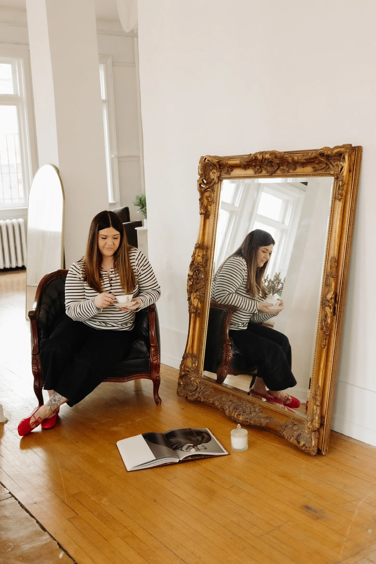 A woman with long brown hair, wearing a striped shirt, black pants, and red shoes, is seated on a vintage armchair in a room with white walls and wooden floors, holding a teacup. A large, ornate gold-framed mirror leaning against the wall reflects her. An open magazine and a lit candle are on the floor nearby.