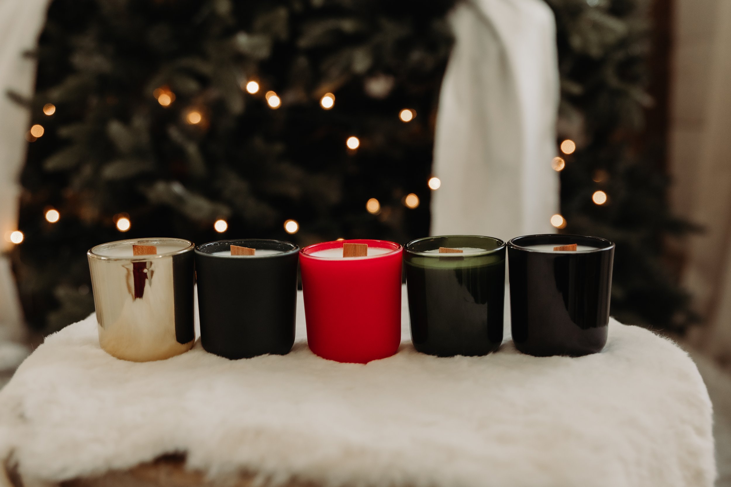 A row of five candles in various colors, placed on a white surface with a blurred Christmas tree with lights in the background.