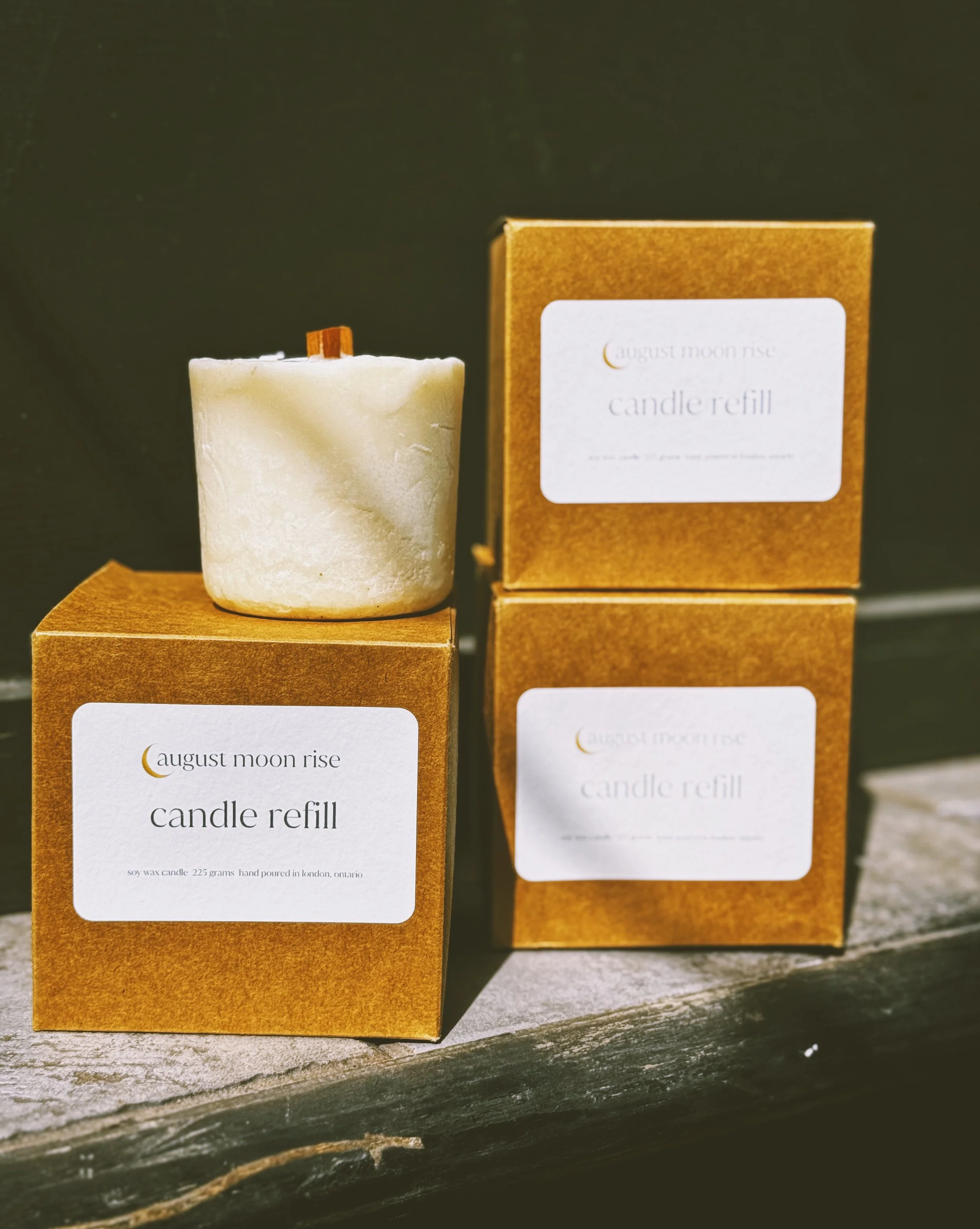 A white candle on top of a gold box labeled 'August Moon Rise candle refill,' with two similar boxes stacked beside it on a wooden surface.