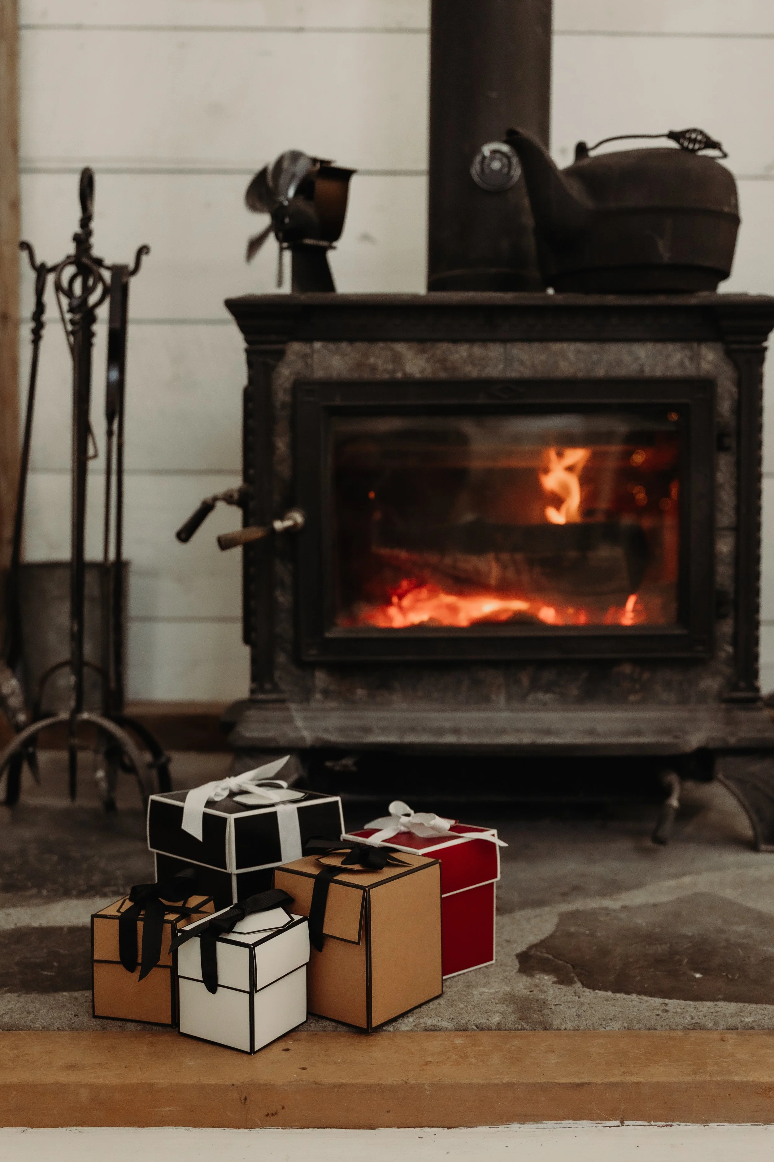 A wood-burning stove with flames inside, situated in a cozy indoor setting. On top of the stove are old-fashioned black cast iron boots. To the left are a set of fireplace tools standing in a holder, and below are wrapped gift boxes with bows, featuring black, white, red, and brown wrapping paper.