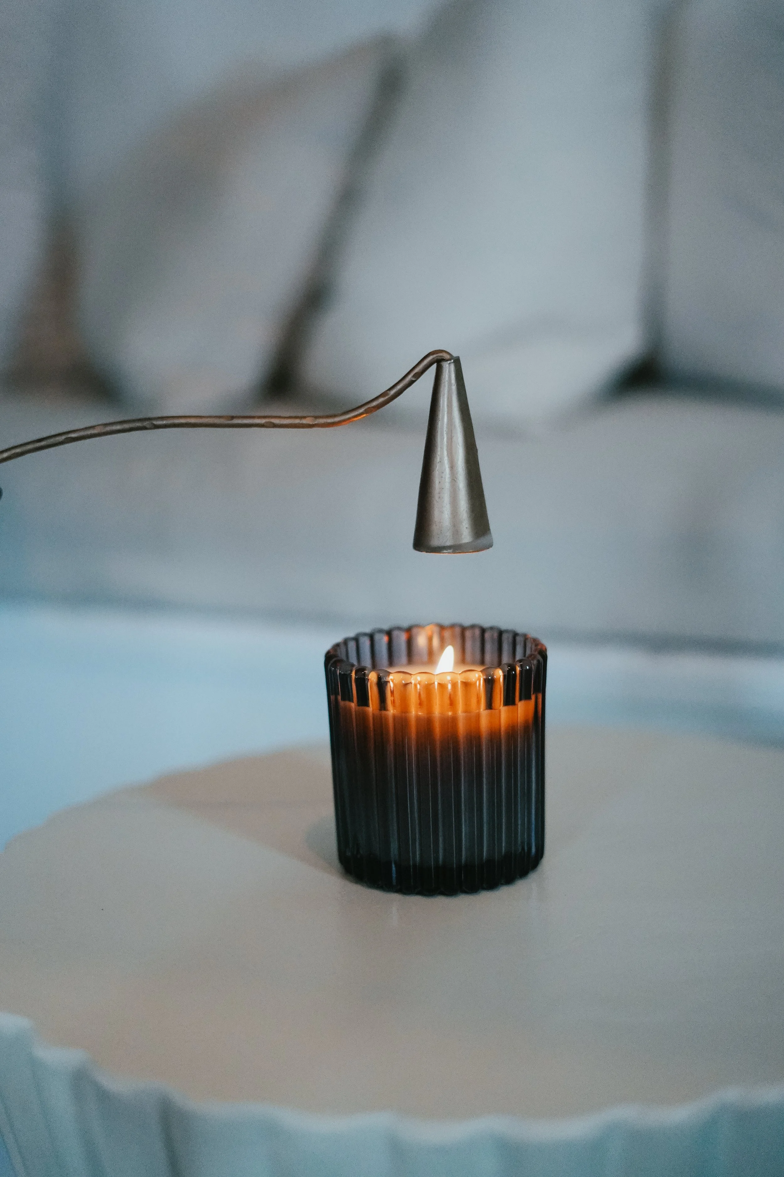 A lit candle in a black ribbed glass holder on a white surface, with a metal candle snuffer above it.