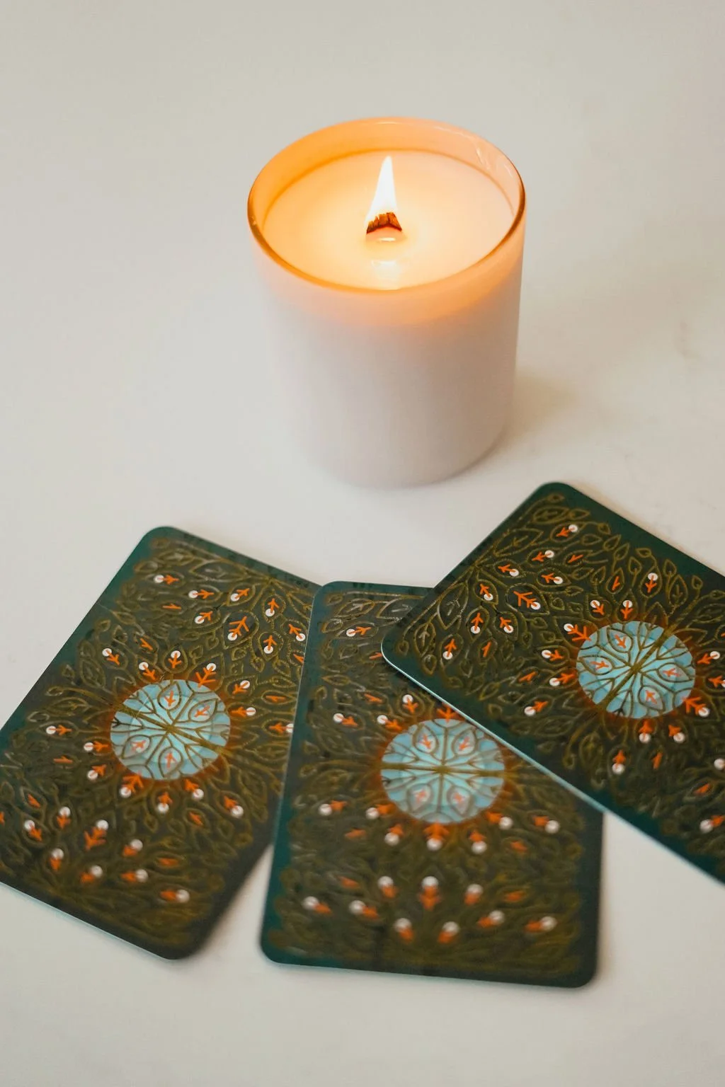 A white candle with a burning flame and three tarot cards with intricate designs and patterns.