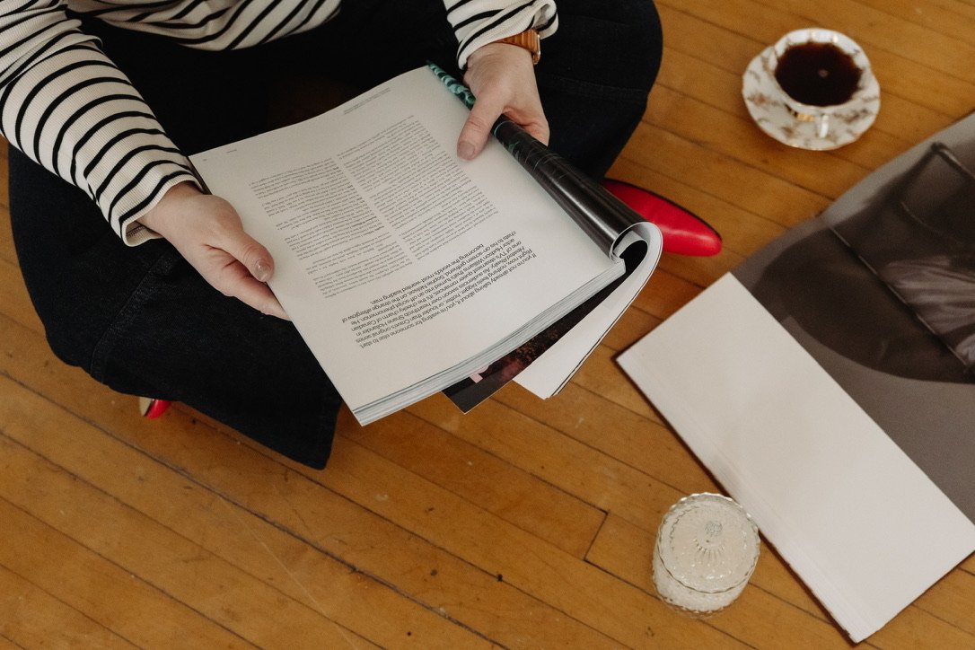 Person in a striped shirt sitting on a red chair, flipping through a large open book with text. Items on the wooden floor include a cup of coffee on a saucer, a small crystal candle holder, and a large bag or rolled-up item.