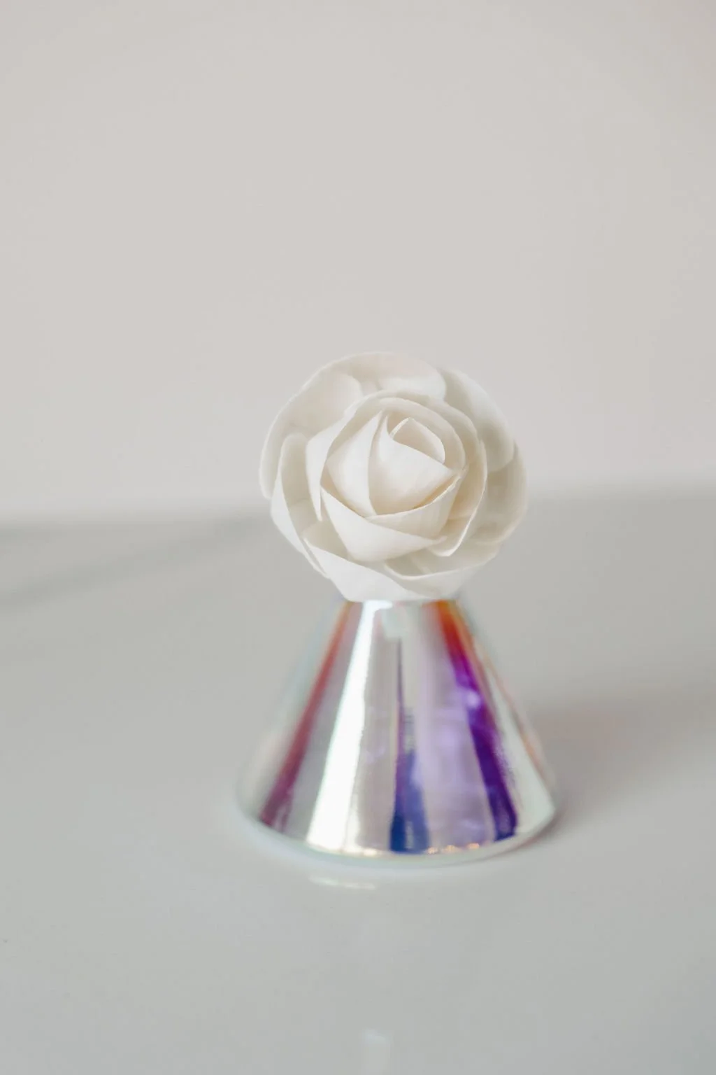 A white rose sola flower placed on top of a small, iridescent cone-shaped diffuser vasewith a reflective surface.