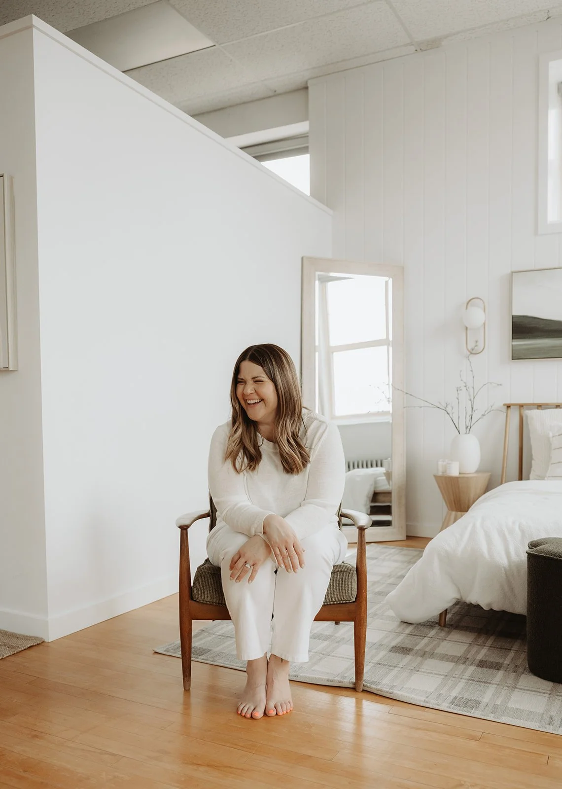 A woman sitting on a chair in a bright, modern bedroom, smiling and laughing.