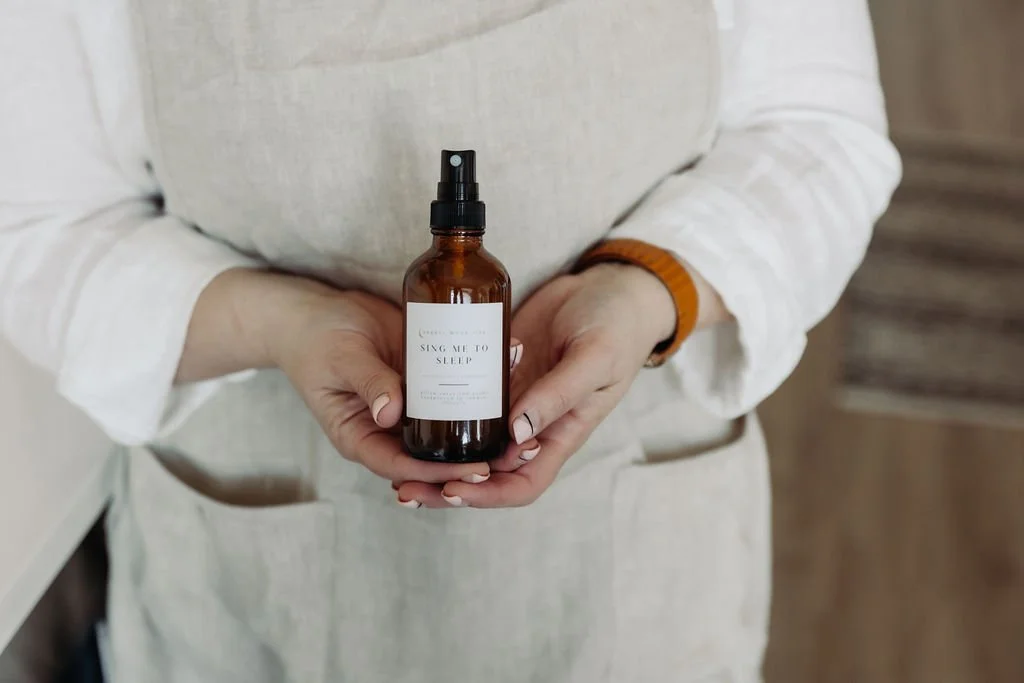 Person holding an amber glass spray bottle labeled "Sing Me to Sleep," wearing a linen apron. The hands display neatly manicured nails, and a wooden floor background is visible.