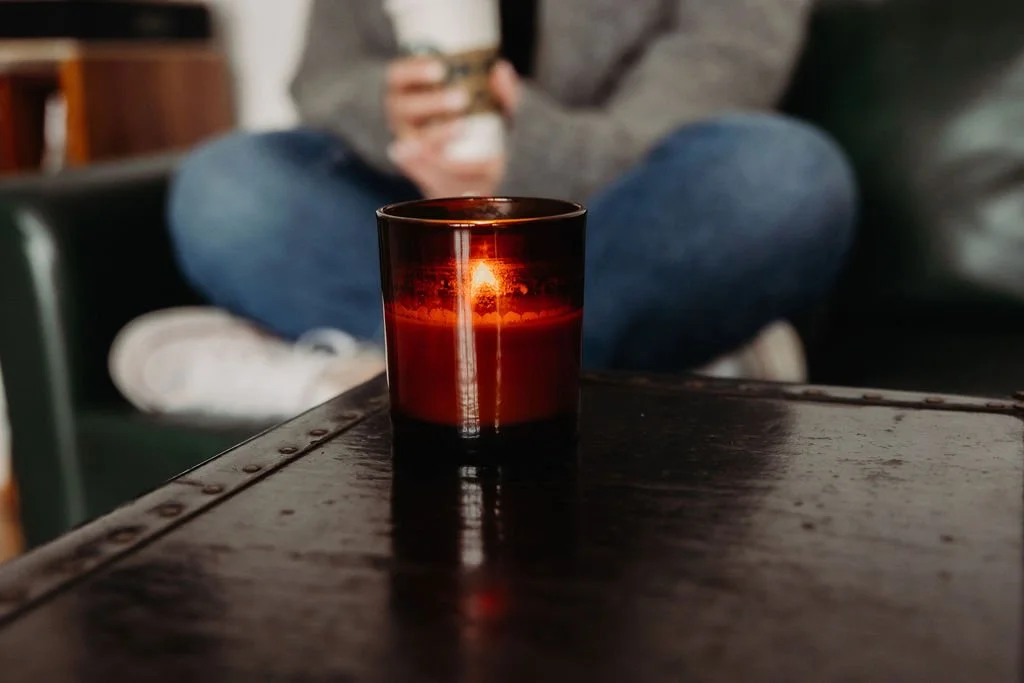 Lit candle in a glass holder on a table, person sitting cross-legged in the background holding a coffee cup.