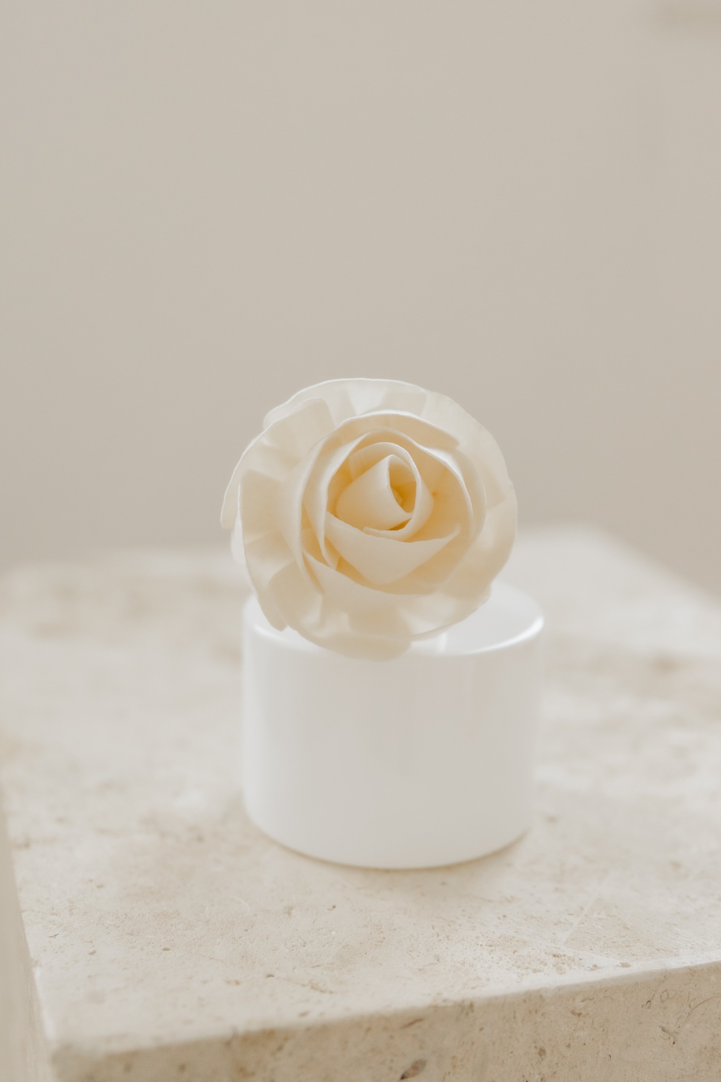 White ceramic vase with a decorative white rose made of a soft material, placed on a white surface against a neutral background.