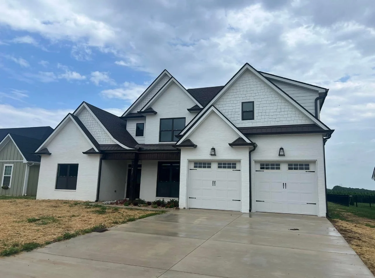 A modern two-story white house with black trim and a two-car garage, situated on a driveway with patches of grass and a partly cloudy sky overhead.