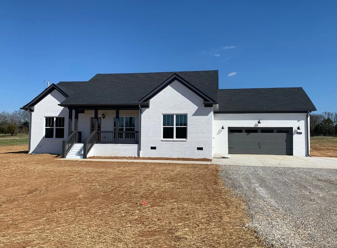 Newly built white house with black roof and trim, front porch with steps, attached two-car garage, and dirt front yard under a clear blue sky.