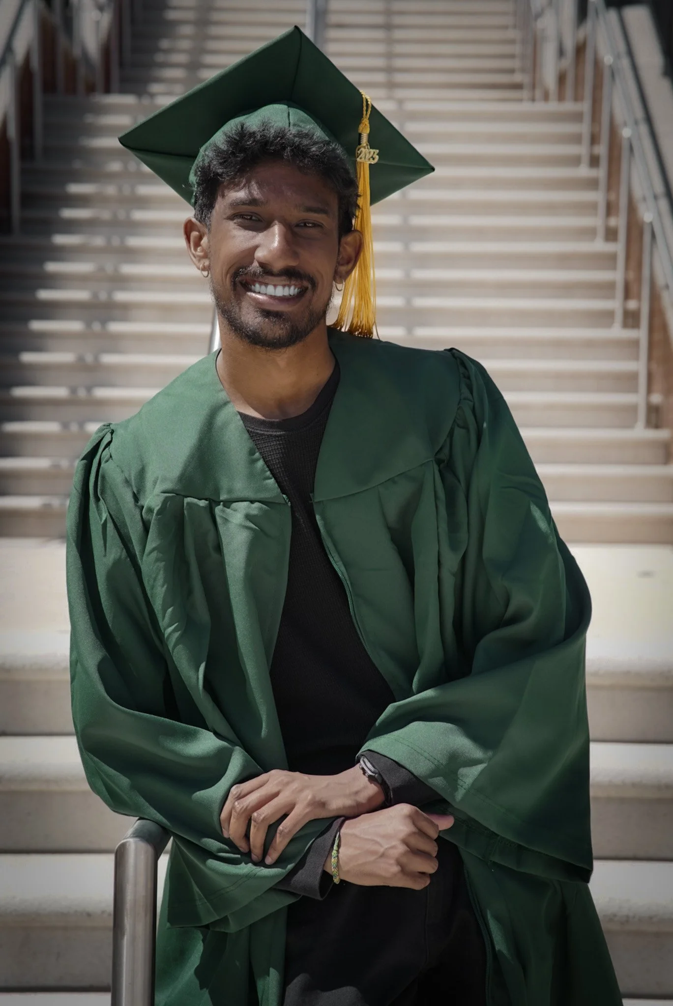 A man in a green graduation gown and cap sitting on outdoor stairs, smiling at the camera.