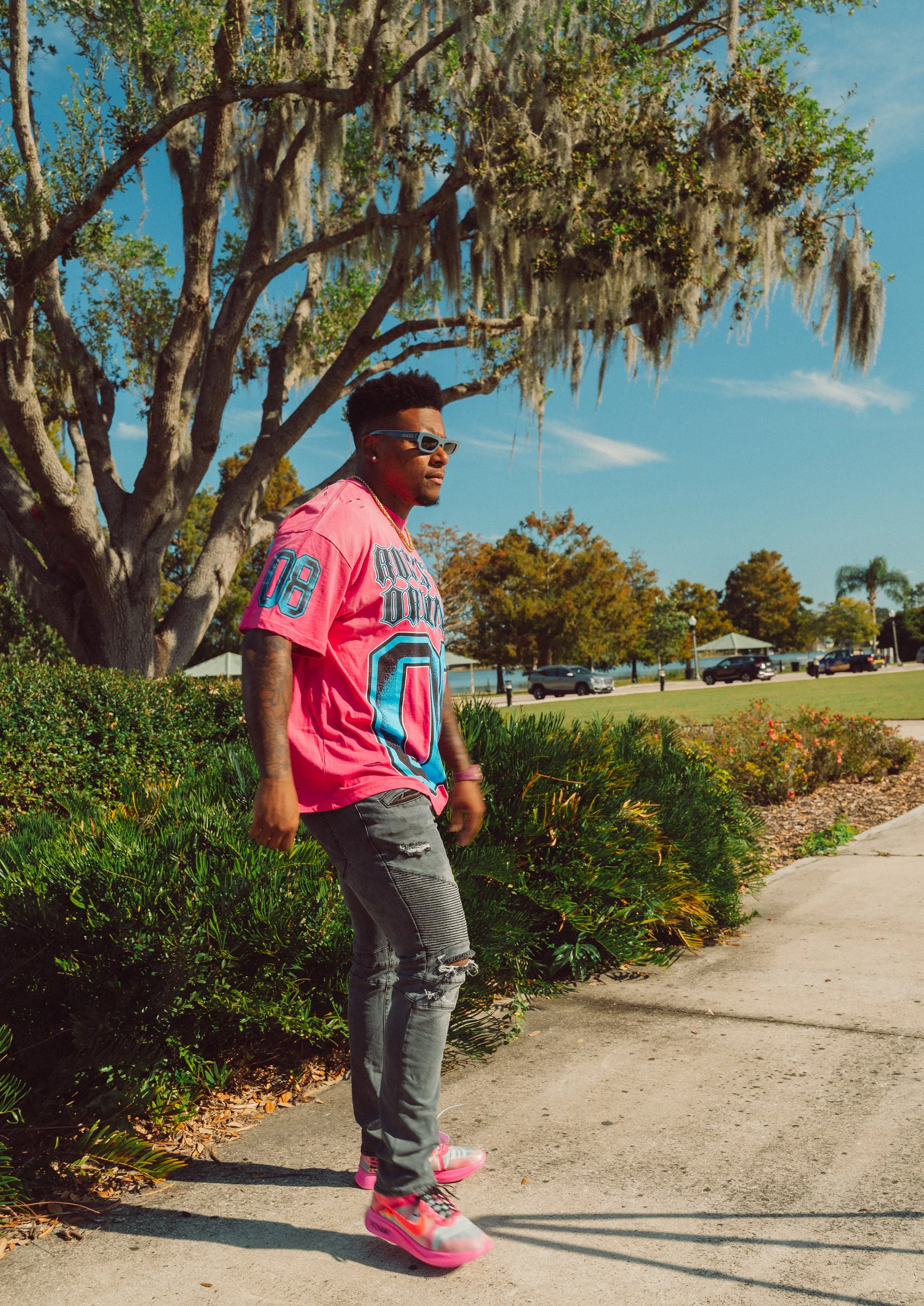 A young man wearing sunglasses, a pink sports jersey, ripped gray jeans, and colorful sneakers is walking outdoors on a sidewalk near green bushes and a large tree with Spanish moss, under a clear blue sky.