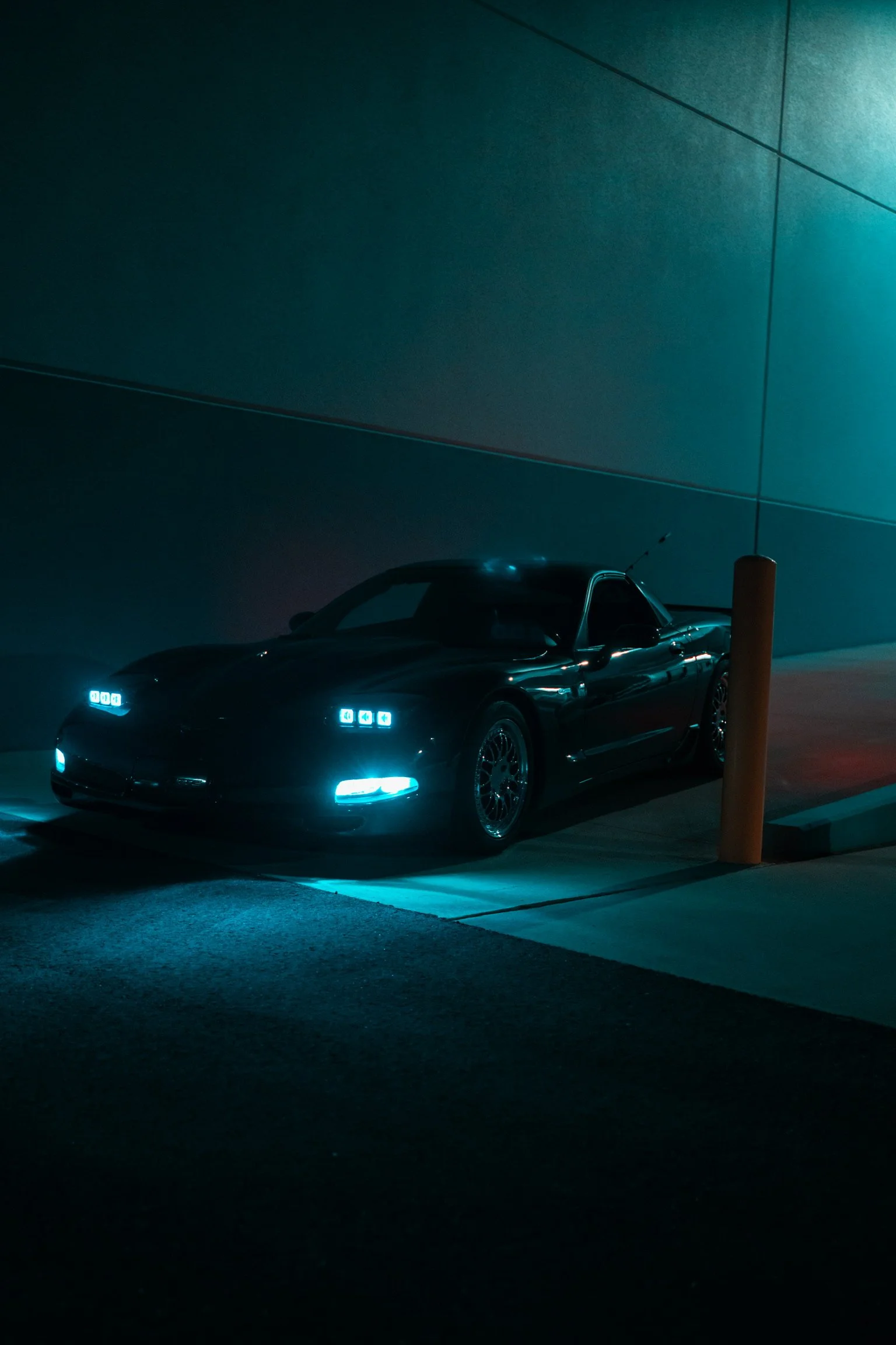 A black sports car parked at night under teal lighting, next to a yellow parking bollard, against a plain teal wall.