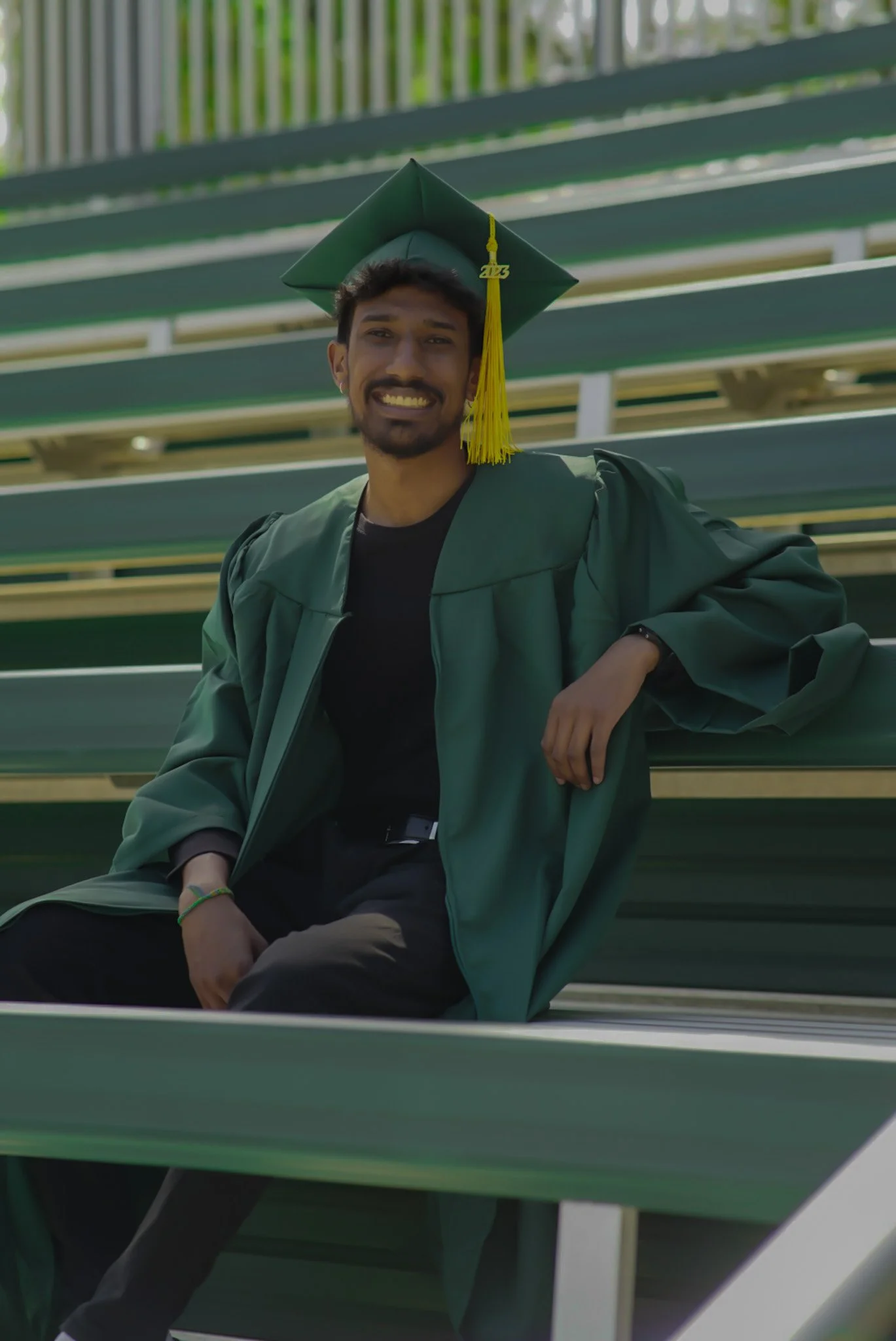 Young man wearing green graduation gown and cap with yellow tassel, sitting on green bleachers, smiling.