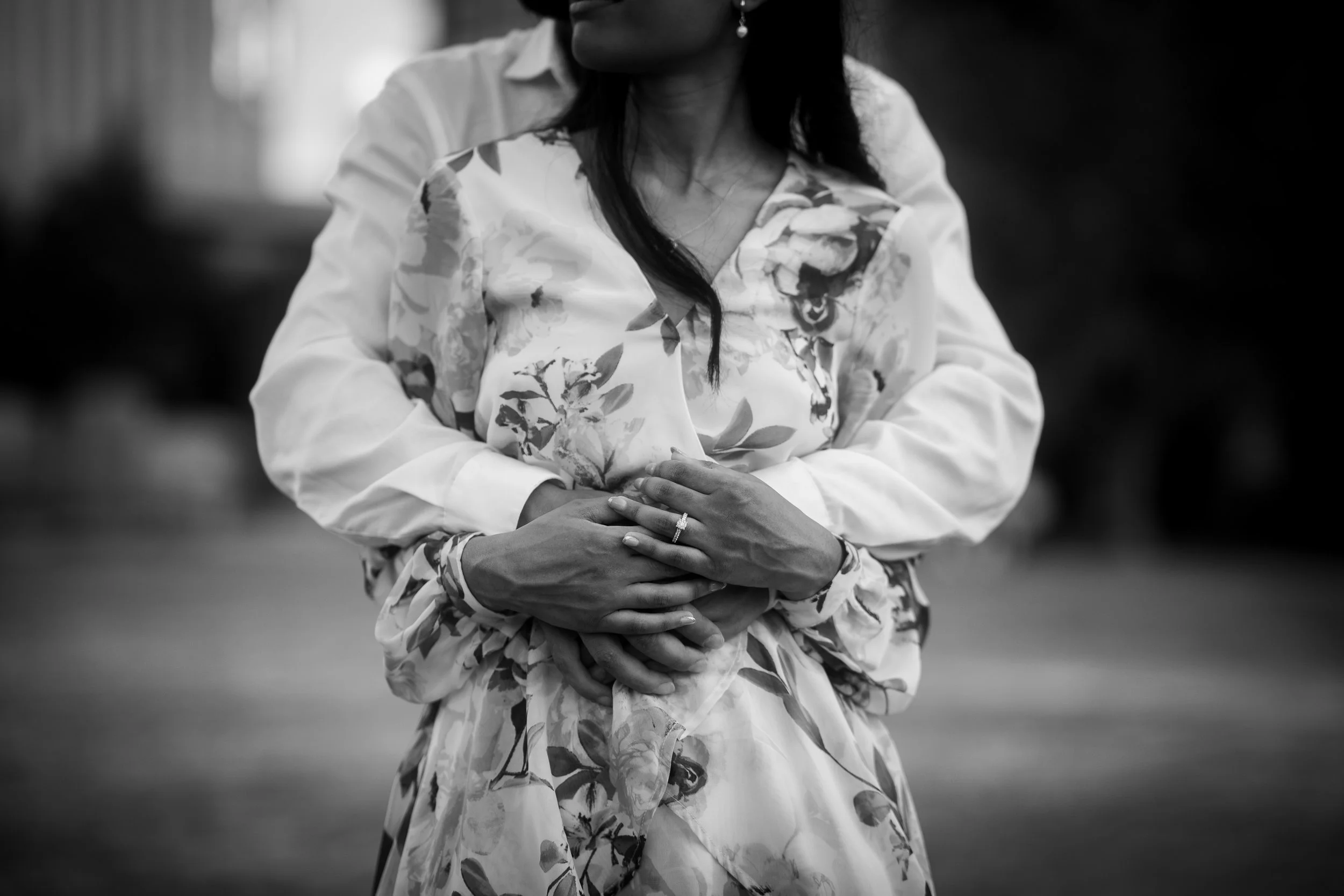 A woman in a floral dress being embraced from behind by another person, both with their hands together at the woman's waist, in an outdoor setting.