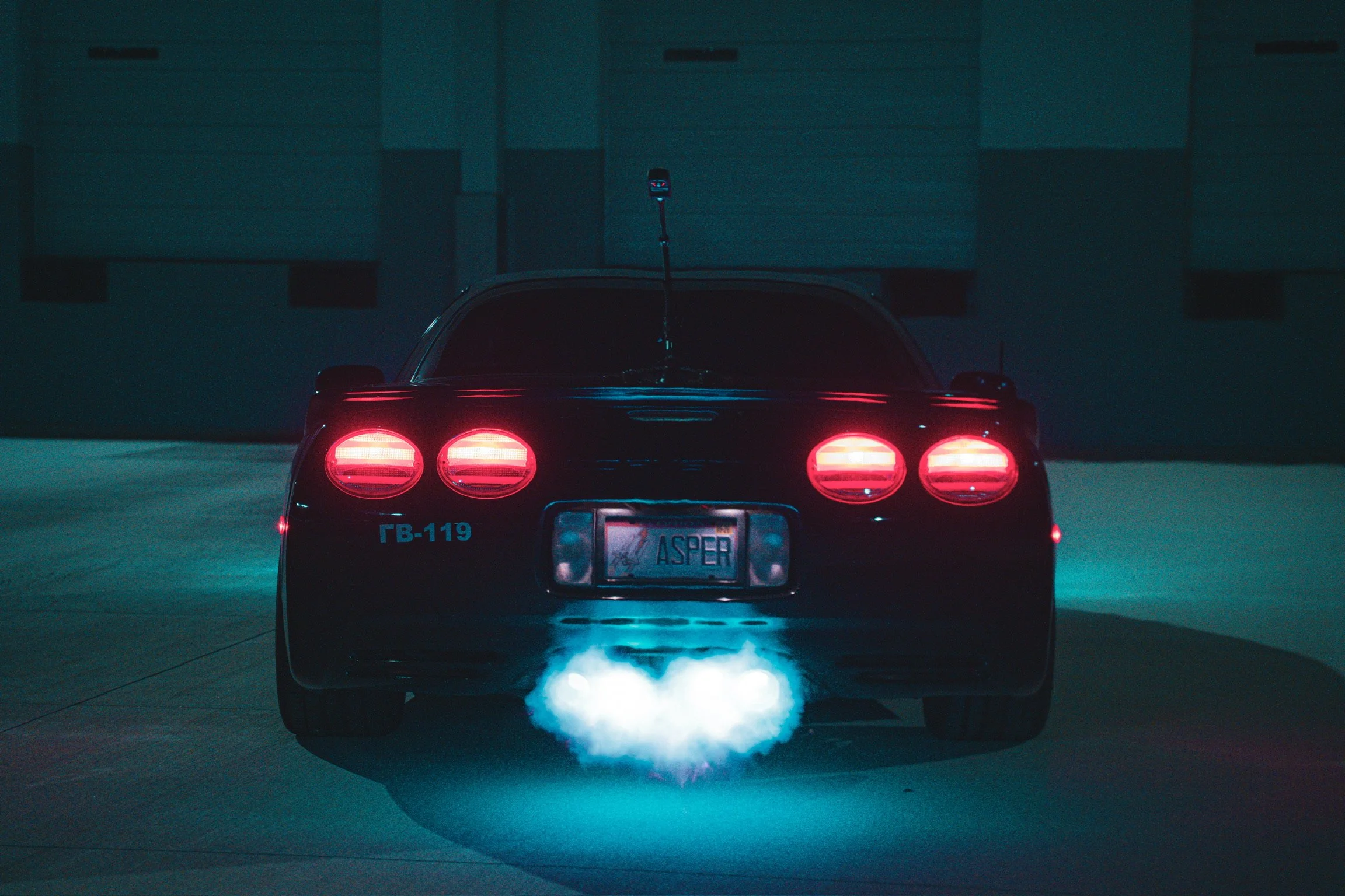A black sports car with red tail lights parked in a dimly lit garage, emitting exhaust fumes in the nighttime.