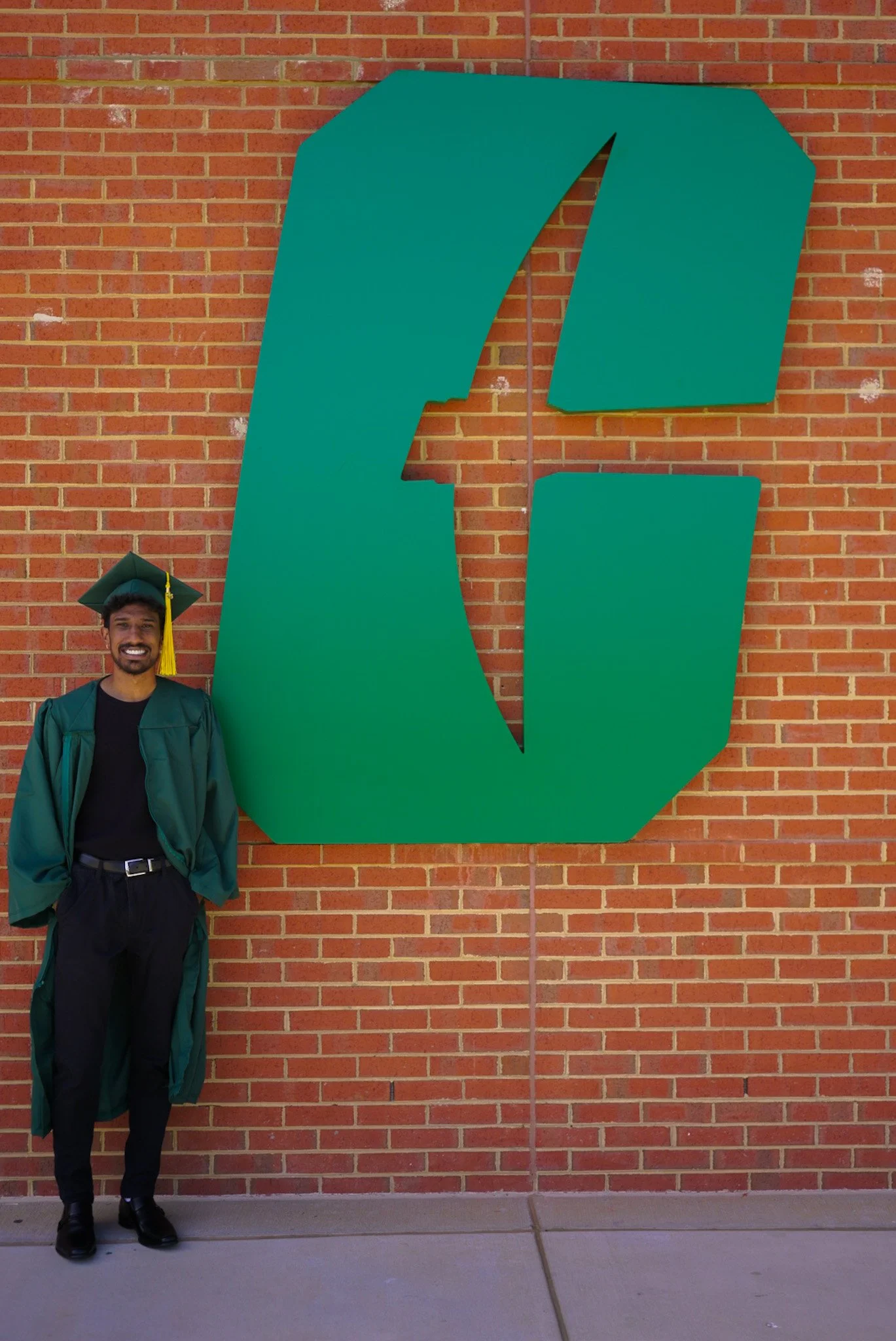 A smiling young man in a graduation cap and gown standing in front of a large green letter 'C' sign on a brick wall.
