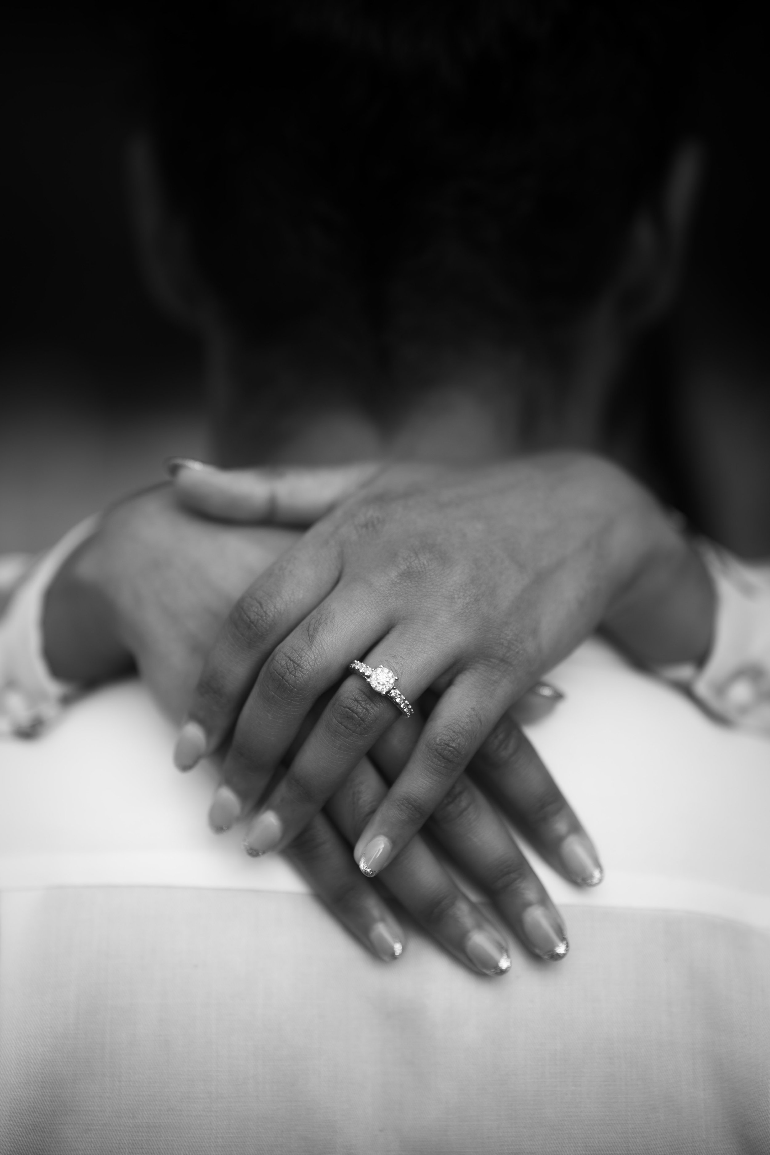 Close-up of a woman's hands with an engagement ring, resting on a surface, with dark hair and face out of focus in the background