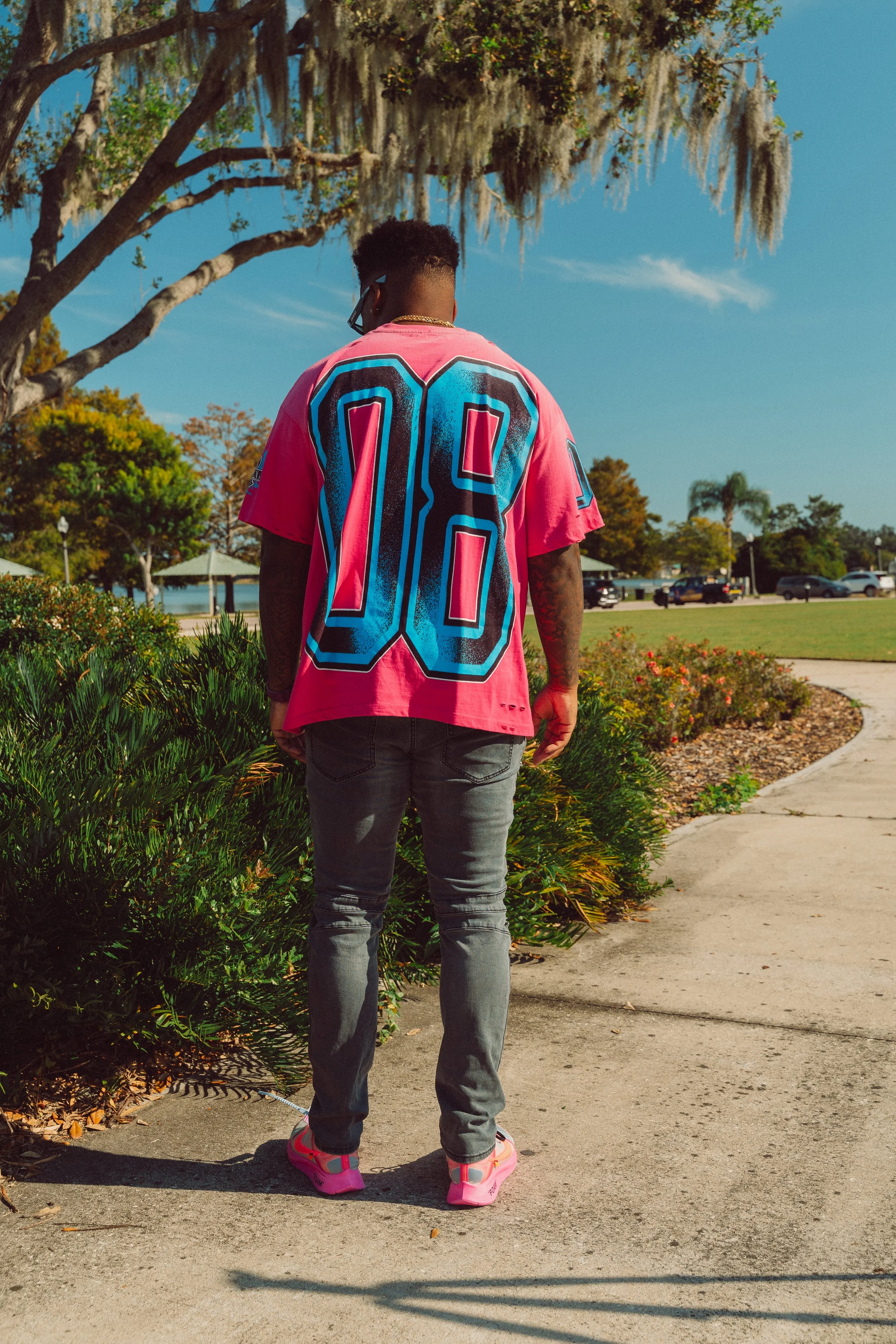 A person standing on a sidewalk near some bushes, wearing a pink sports jersey with the number 02 on the back, gray jeans, pink sneakers, and sunglasses, with trees and blue sky in the background.