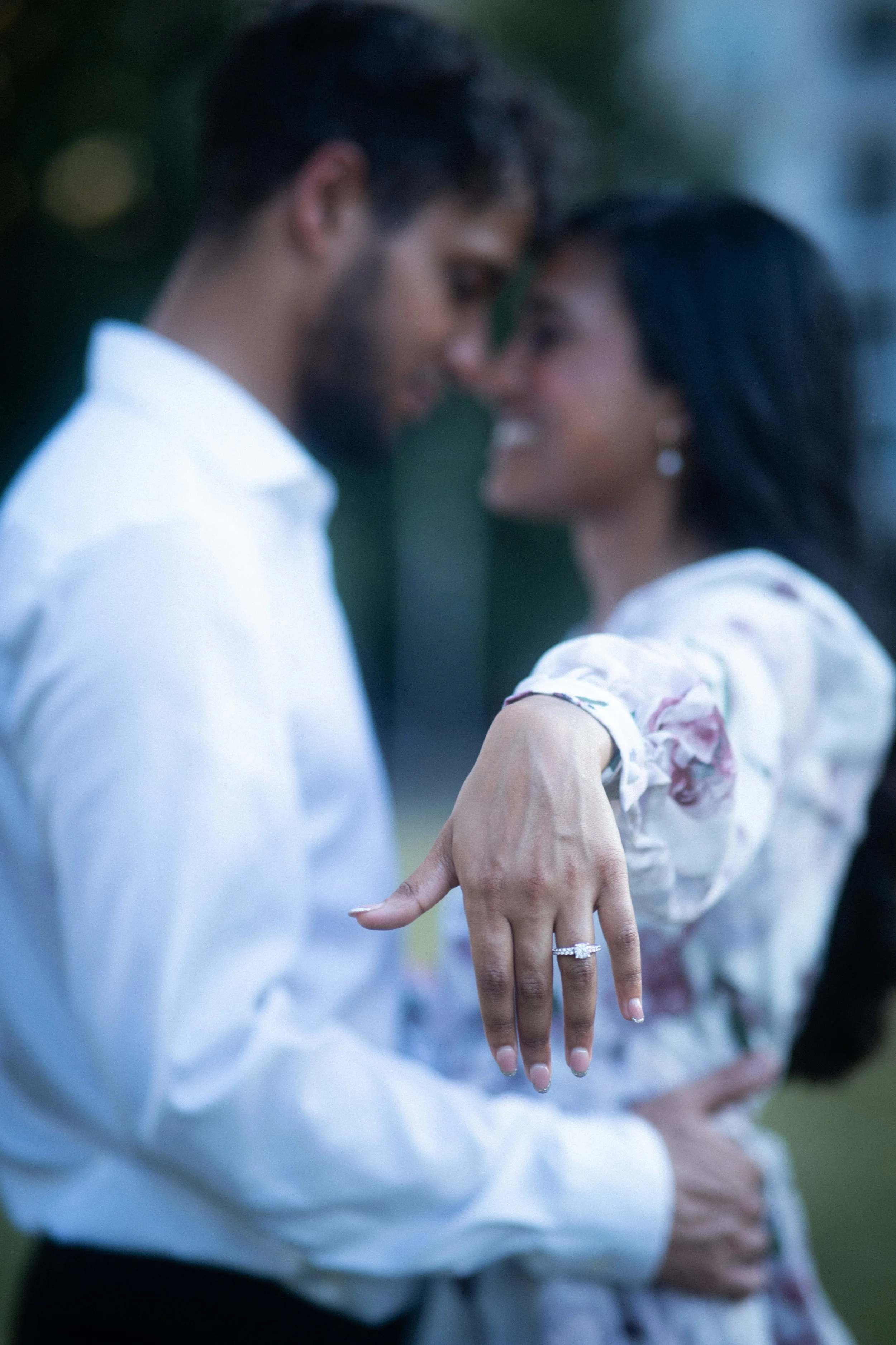 A couple stands close together with their foreheads touching, smiling, and holding hands. The woman is showing off an engagement ring on her finger.