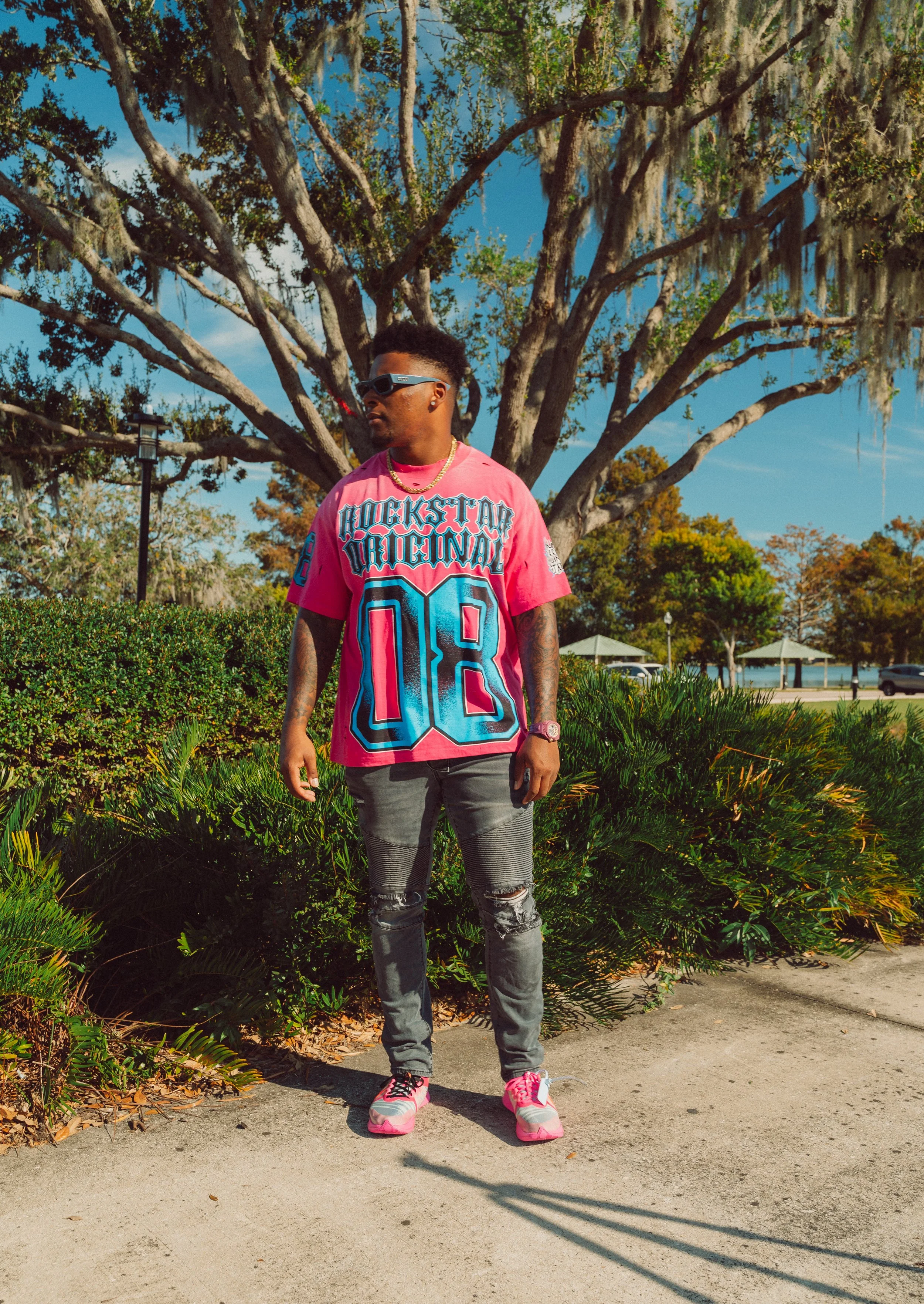 A young man wearing a pink graphic T-shirt, distressed jeans, pink and white sneakers, sunglasses, and a gold chain, standing outdoors on a sidewalk with green bushes and trees behind him. The sky is clear and blue.