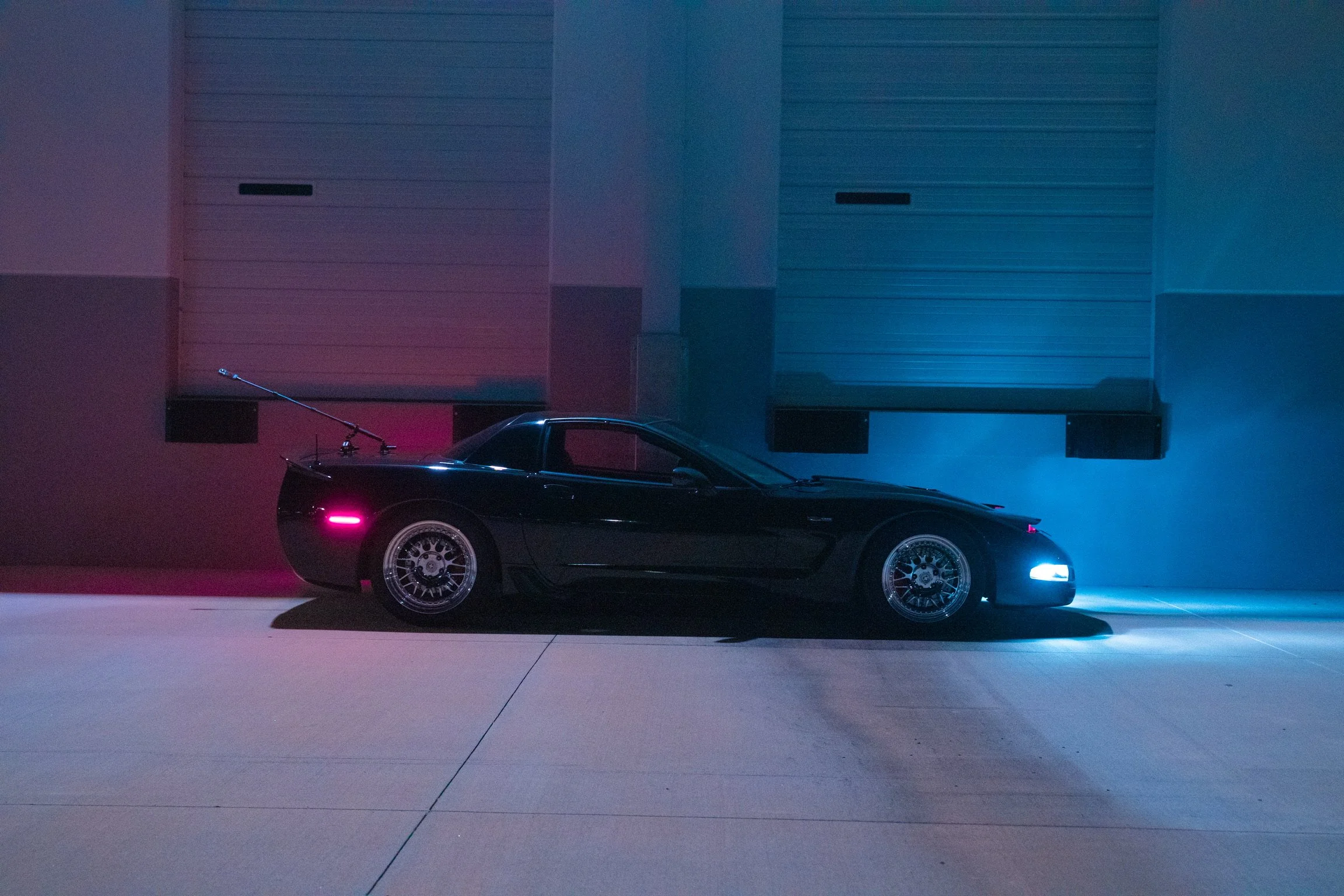 A sleek black sports car with glowing headlights parked in front of a modern industrial building illuminated with colorful neon lights, creating a vibrant and stylish nighttime scene.