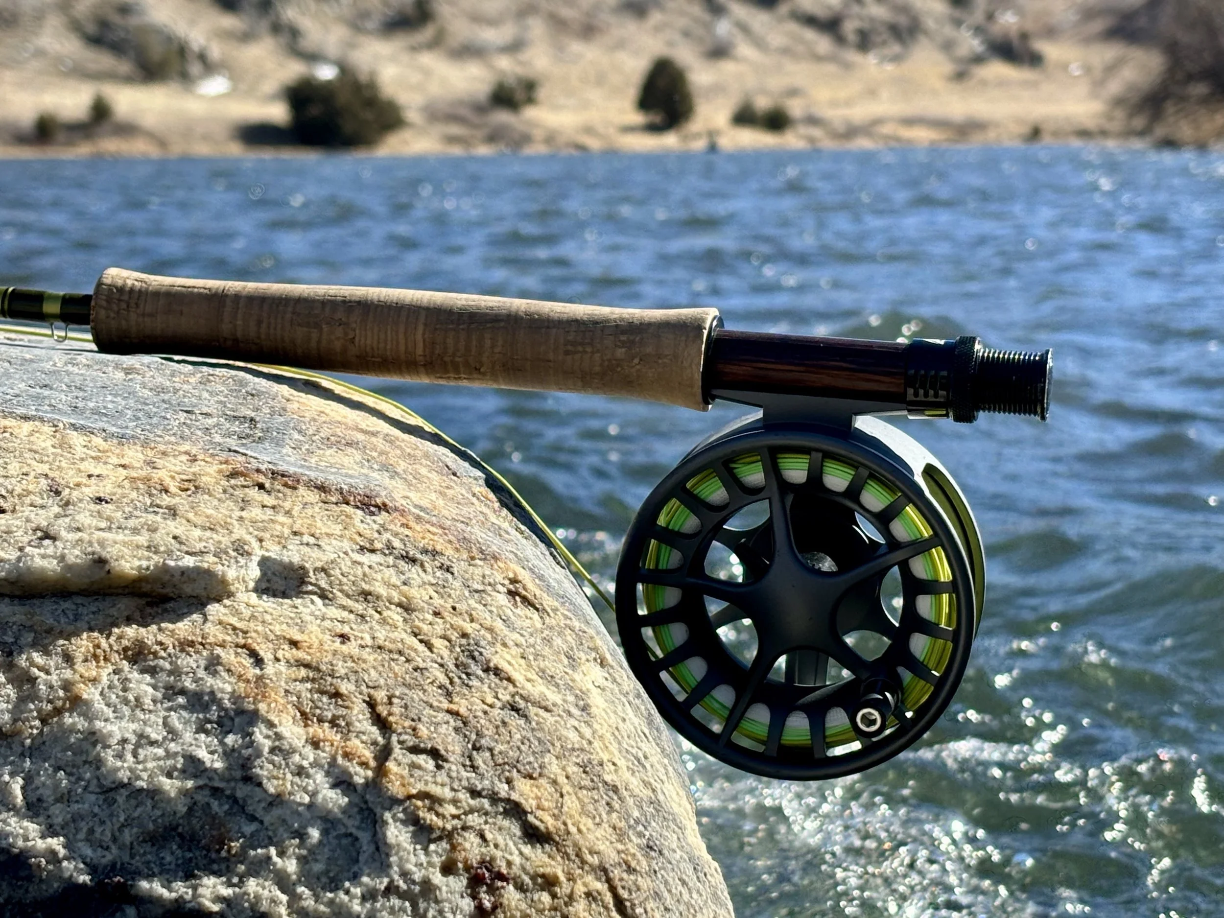 Fly fishing rod and reel resting on a rock by a lake with a blurred background.