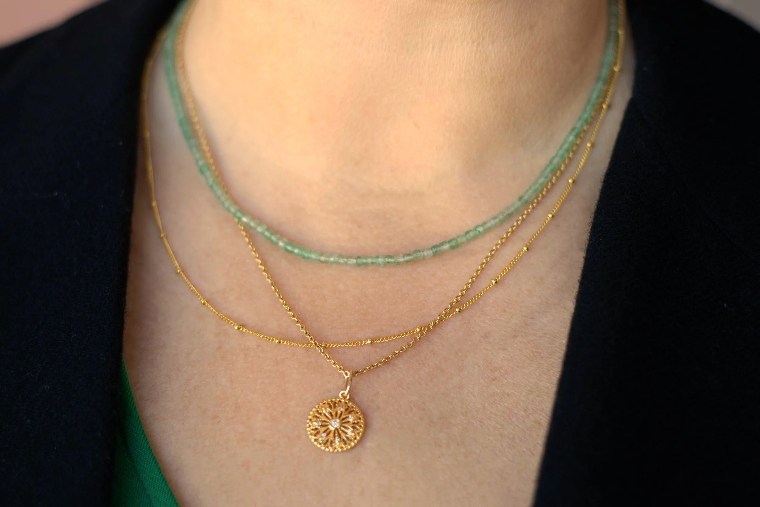 Multiple layered gold necklaces, one with a circular, intricate pendant with a small stone in the center, worn over a green top and black blazer.