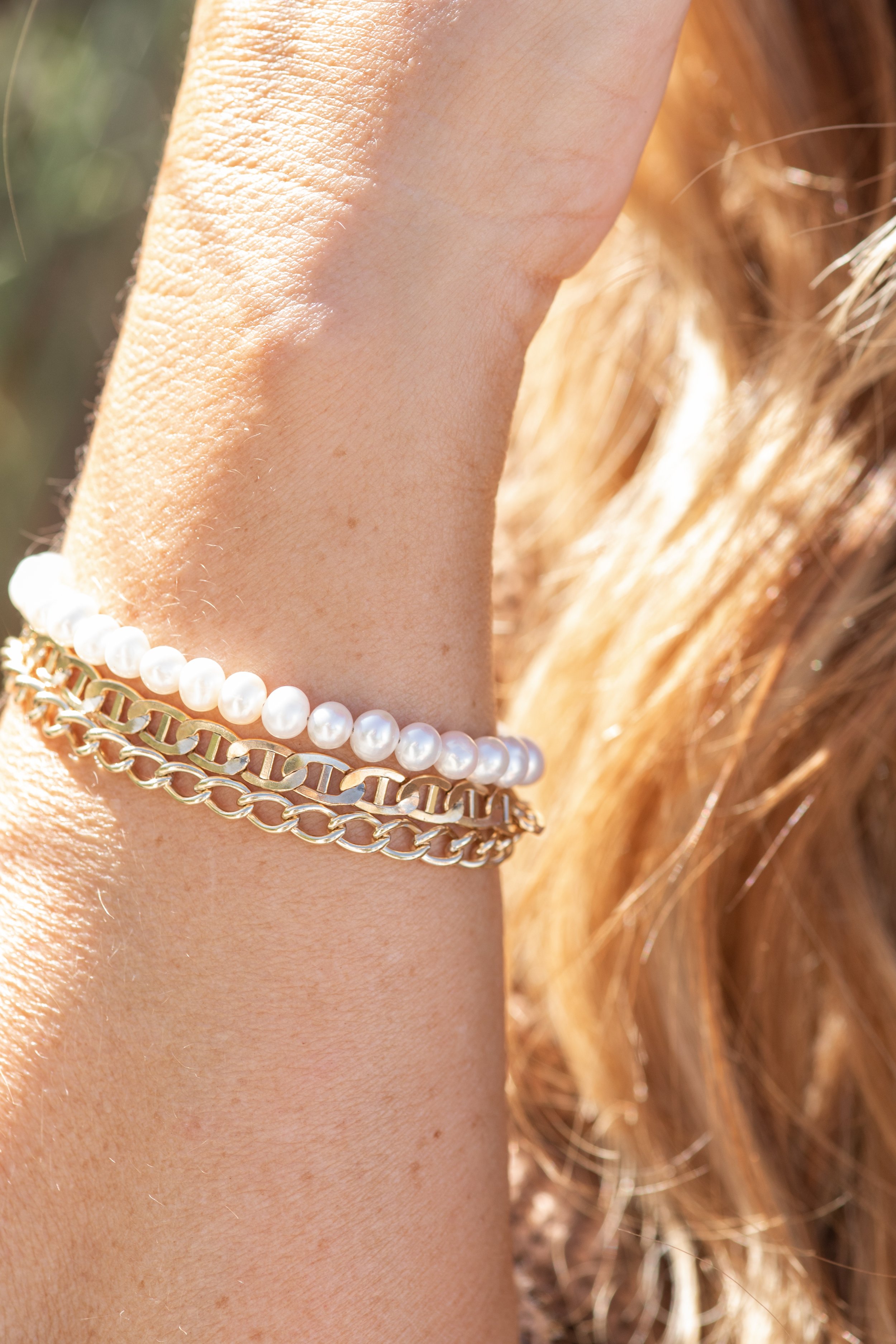 Close-up of a woman's arm wearing three bracelets: a string of white pearls, a gold chain, and a gold link bracelet. The background is blurred and the woman has long, wavy, blonde hair.