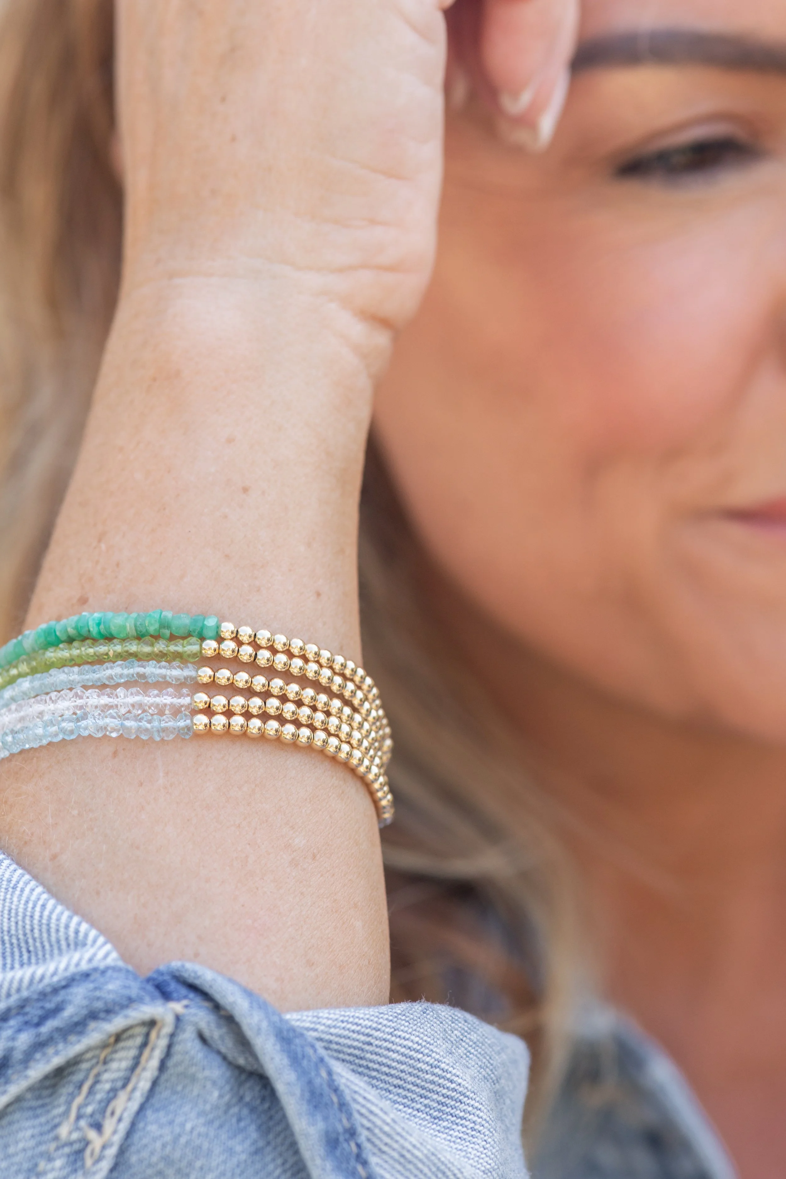 Close-up of a woman's face and wrist, showing her wearing multiple beaded bracelets in green, blue, and gold. She is gently touching her ear with her hand.