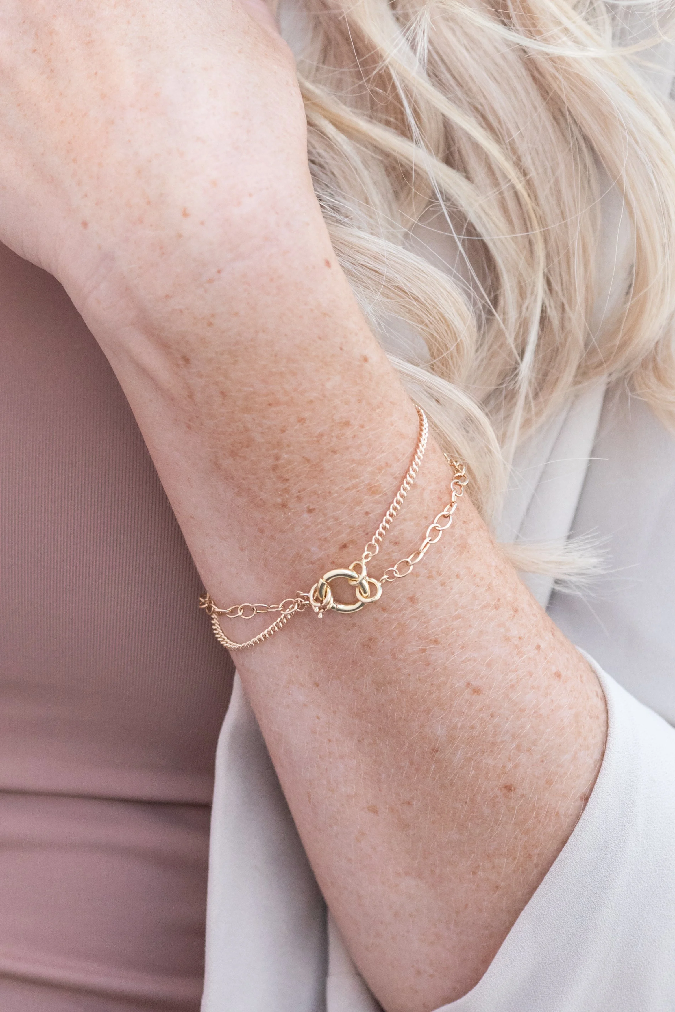 Close-up of a woman's arm with a gold chain bracelet, blonde hair, and wearing a light-colored sleeve and a mauve top.