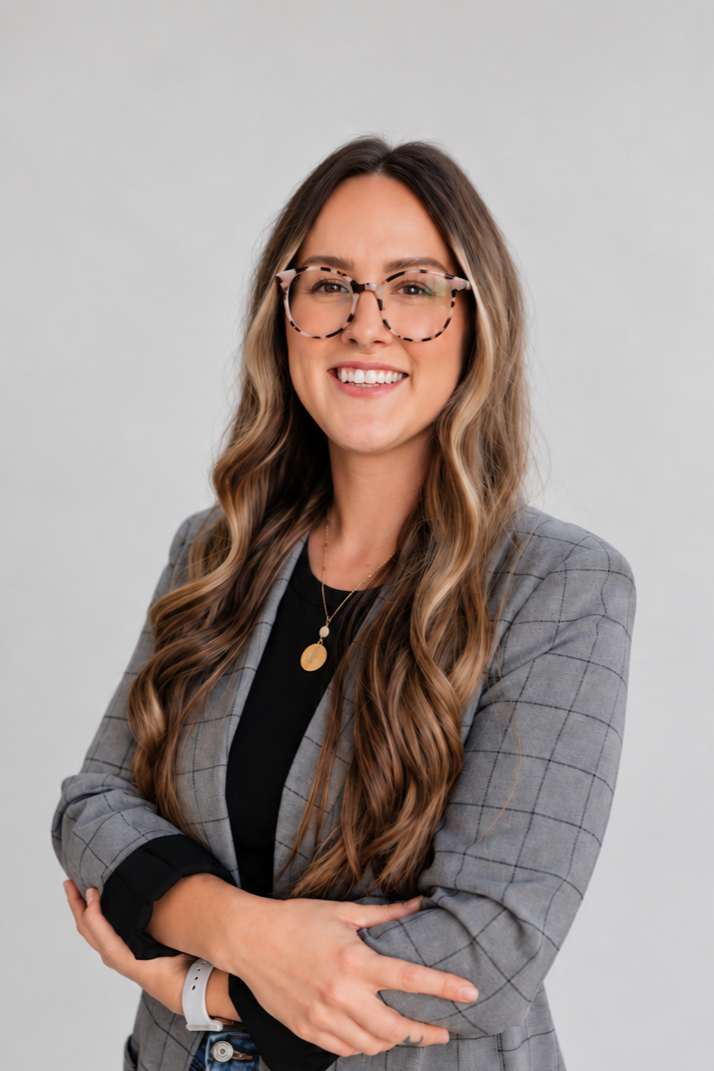 A woman with long wavy blonde hair, wearing glasses with a tortoiseshell pattern, a grey checkered blazer over a black top, and a gold necklace, smiling and standing against a plain white wall.