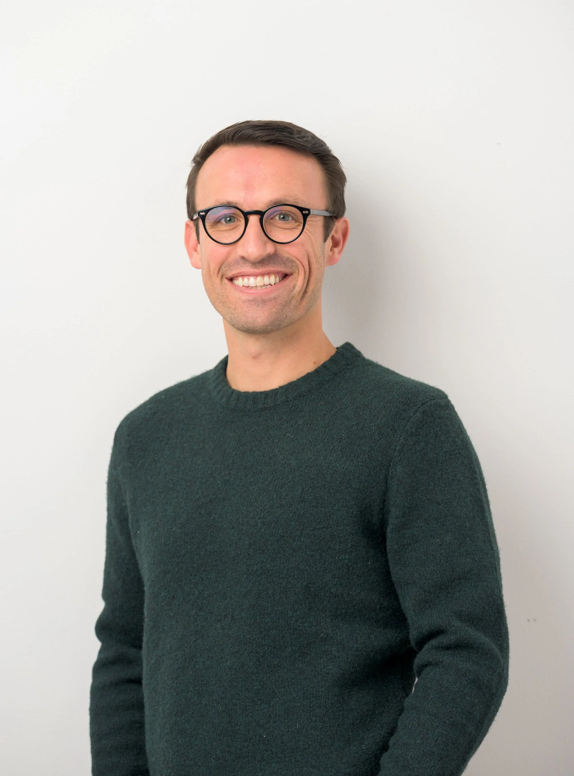 A smiling woman with shoulder-length blonde hair, wearing a black turtleneck and a patterned blazer, standing against a plain white wall.