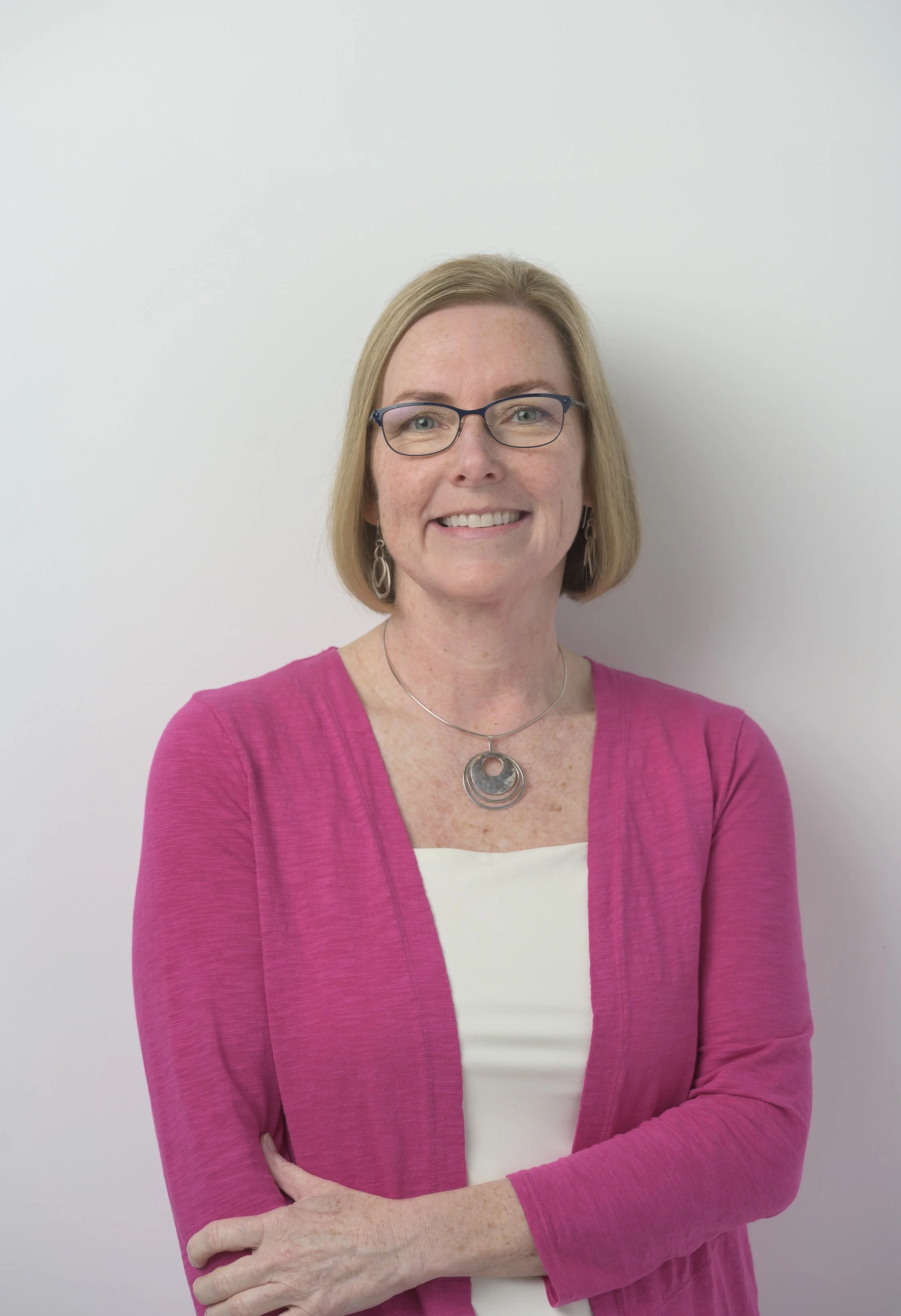 A woman with blonde hair wearing glasses, a black cardigan, and a black and white patterned top, smiling in front of a plain white background.