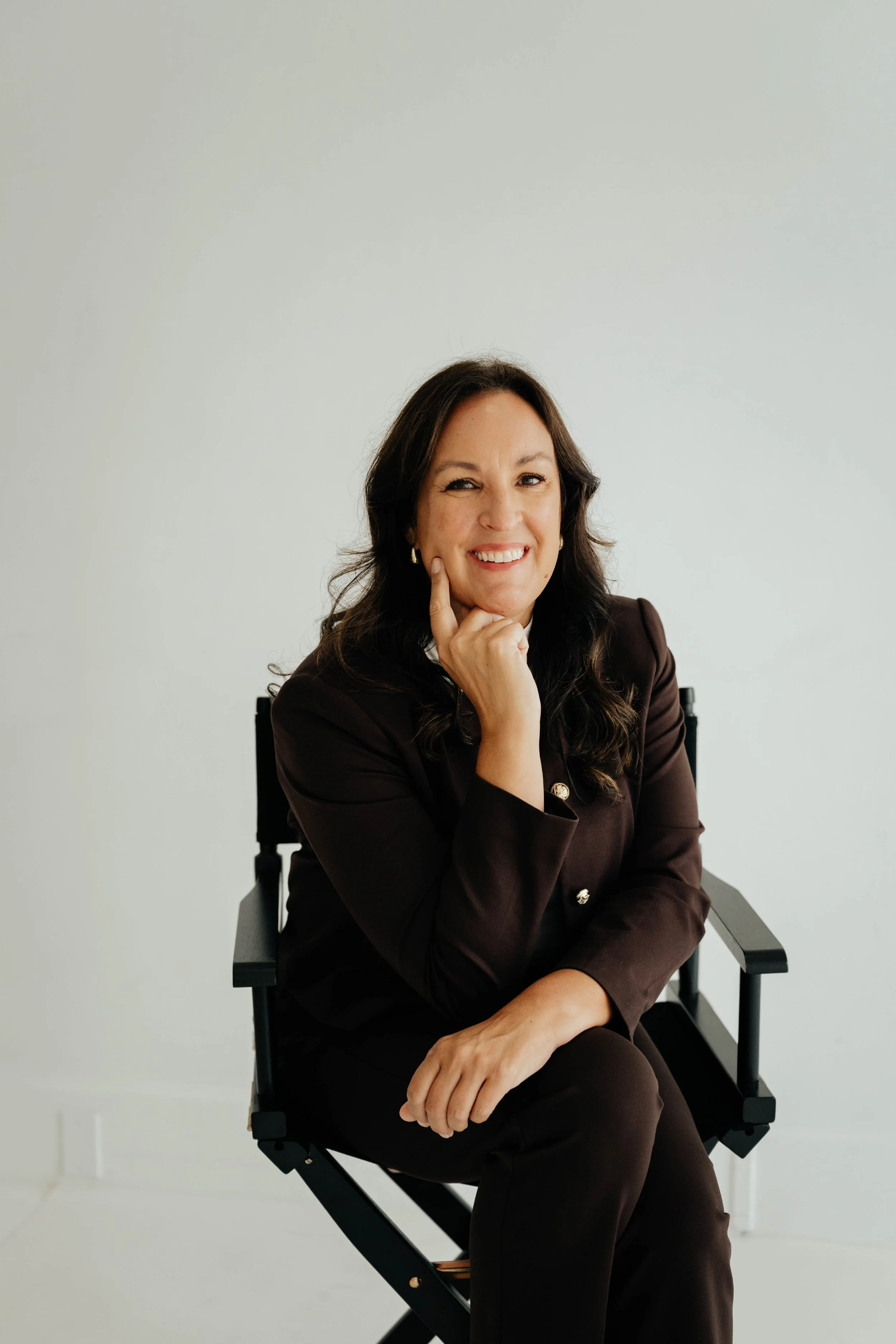 A woman with shoulder-length dark hair smiling, wearing a purple long-sleeve top, gold hoop earrings, and a gold necklace, against a plain white background.